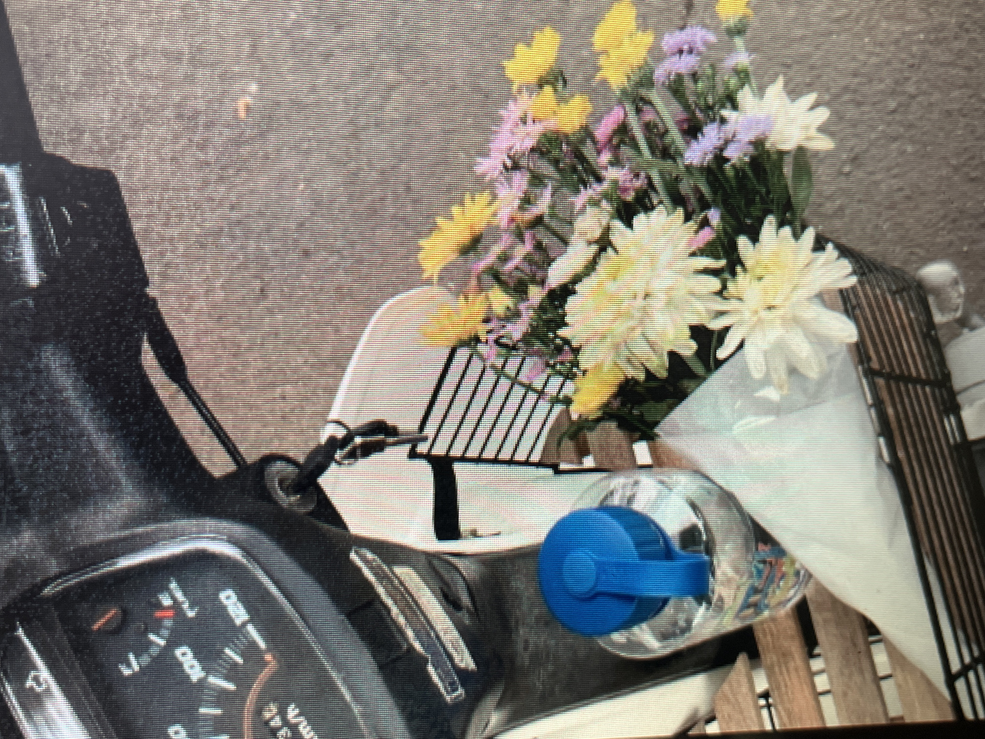 The image shows a close-up view of a scooter's handlebar area with a basket attached. In the basket, there is a clear plastic water bottle with a blue lid, and a bouquet of mixed flowers, predominantly yellow and purple, with some white blooms. The scooter's dashboard with a speedometer is visible on the left side of the frame. The background appears to be a paved surface, possibly a street or sidewalk. There are no people or any activity depicted in the image. The scene is static and focuses on the objects in the scooter's basket and dashboard. The location is not specified in the image itself.
