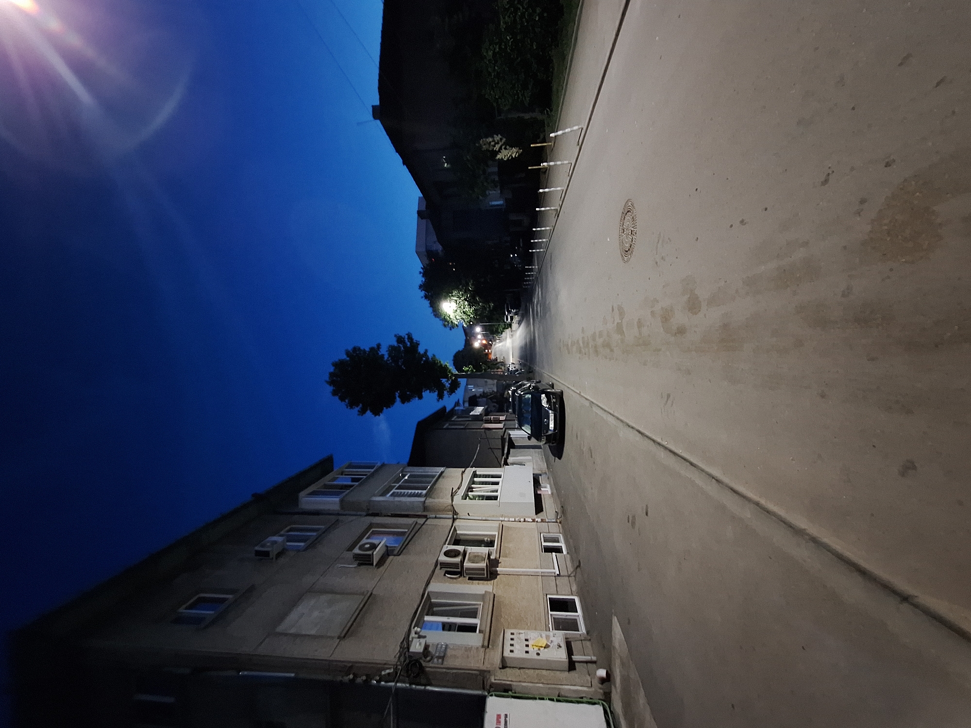 The image depicts a street scene at dusk in Varna, Bulgaria. The sky is a deep, clear blue, transitioning to black as night falls. A bright lens flare from a light source is visible in the upper right corner. On the left side of the street, a multi-story apartment building with visible air conditioning units and windows is present. A black car is parked on the street. The street itself is paved and appears to be well-lit by streetlights, some of which are visible in the distance. Along the right side of the street, smaller buildings and trees line the road. The overall atmosphere is quiet and urban, illuminated by artificial lights against the twilight sky.