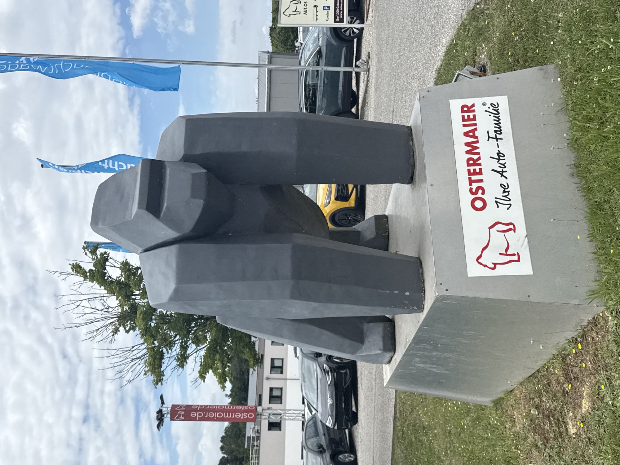 The image shows a large, gray, polygonal sculpture of a gorilla standing on a metallic pedestal. A sign on the pedestal reads "OSTERMAIER Ihre Auto-Familie" with a red outline of a gorilla. Behind the sculpture, a car dealership lot is visible with several cars, including a black Volkswagen ID.3, a yellow car, and a dark gray SUV. In the background, there are buildings, trees, and a cloudy sky. A tall red sign with the text "ostermaier.de" is visible to the left of the sculpture. Blue flags with text are flying in the background. The scene is located in Vilsbiburg, Germany, at the Ostermaier car dealership.