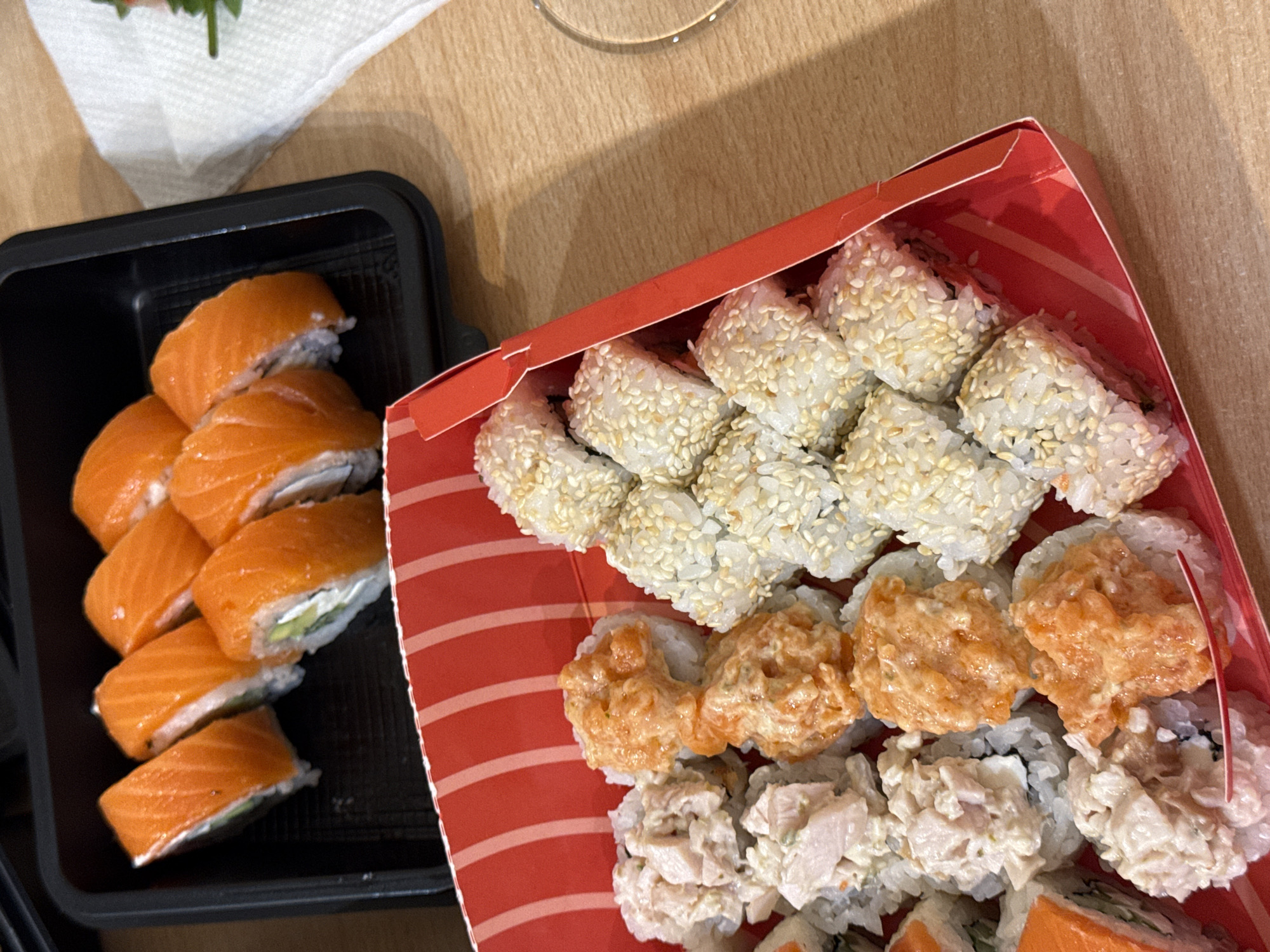 The image shows two containers of sushi on a light brown wooden surface. A black rectangular container on the left contains salmon sushi rolls. A red-and-white striped rectangular container on the right is filled with three rows of sushi: a top row of sesame-seed-covered rolls, a middle row of orange-topped rolls, and a bottom row of white-topped rolls. A white napkin with green vegetation is in the upper left corner. The city and country of origin are Kazan, Russia.