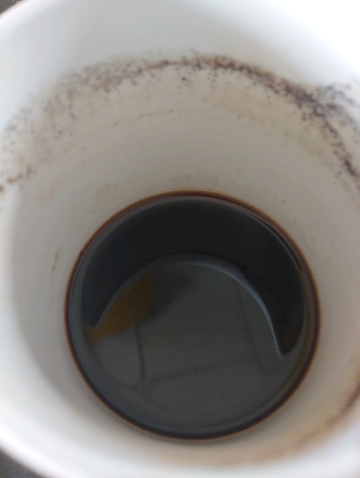 A close-up, overhead shot shows the inside of a white paper cup with a dark liquid, likely coffee, at the bottom. The upper rim of the cup is visible, stained with brown residue, suggesting it has been used. The lighting in the image creates a slight glare on the surface of the liquid, obscuring any fine details of the contents. The background is indistinct, providing no environmental context. There are no people or specific actions depicted. The image is devoid of any geographical markers, and therefore, no information can be provided about its location in Jepara, Indonesia.