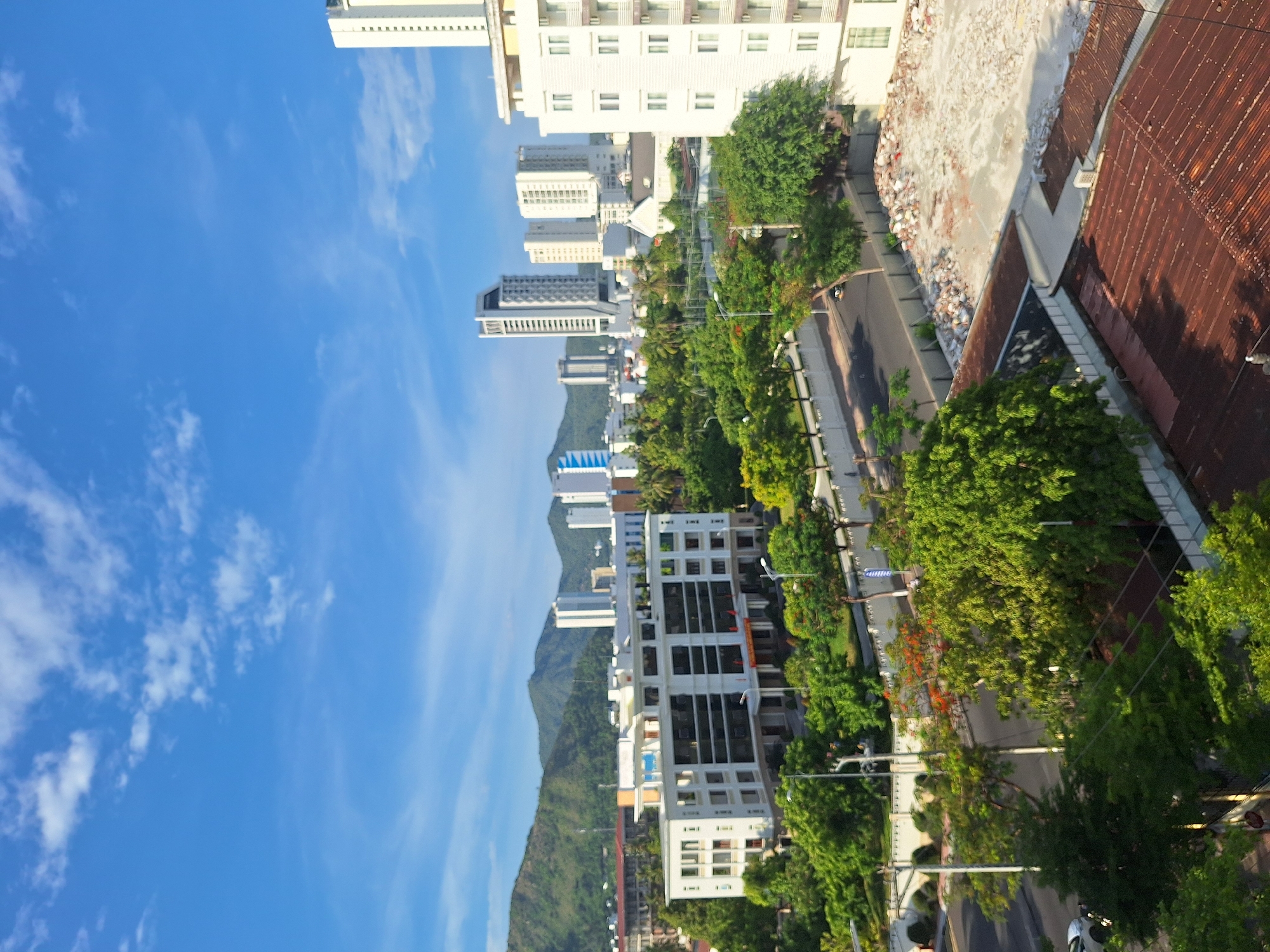 This is a high-angle, vertical shot of a city street in Nha Trang, Vietnam, bathed in the warm light of what appears to be late afternoon or early morning. The sky is a vibrant blue, with wisps of white clouds scattered across it. Dominating the left side of the frame are several modern white buildings of varying heights, some appearing to be residential or hotels, with numerous windows and balconies. In the background, a lush green mountain rises, partially obscured by the buildings.
The right side of the image reveals a street lined with abundant green trees, their leaves catching the sunlight. A road runs alongside the trees, with a few cars visible. There are also streetlights and utility poles. One building in the foreground on the right has a distinctly different roof, appearing to be made of corrugated metal and showing signs of age. Adjacent to this is another structure with a more modern, flat roof, piled with rubble, suggesting construction or demolition activity.
The scene conveys a sense of urban life integrated with nature, typical of coastal cities. No people are clearly visible. The overall mood is peaceful and bright, highlighting the architectural diversity and the verdant surroundings. No visible text is discernable in the image.