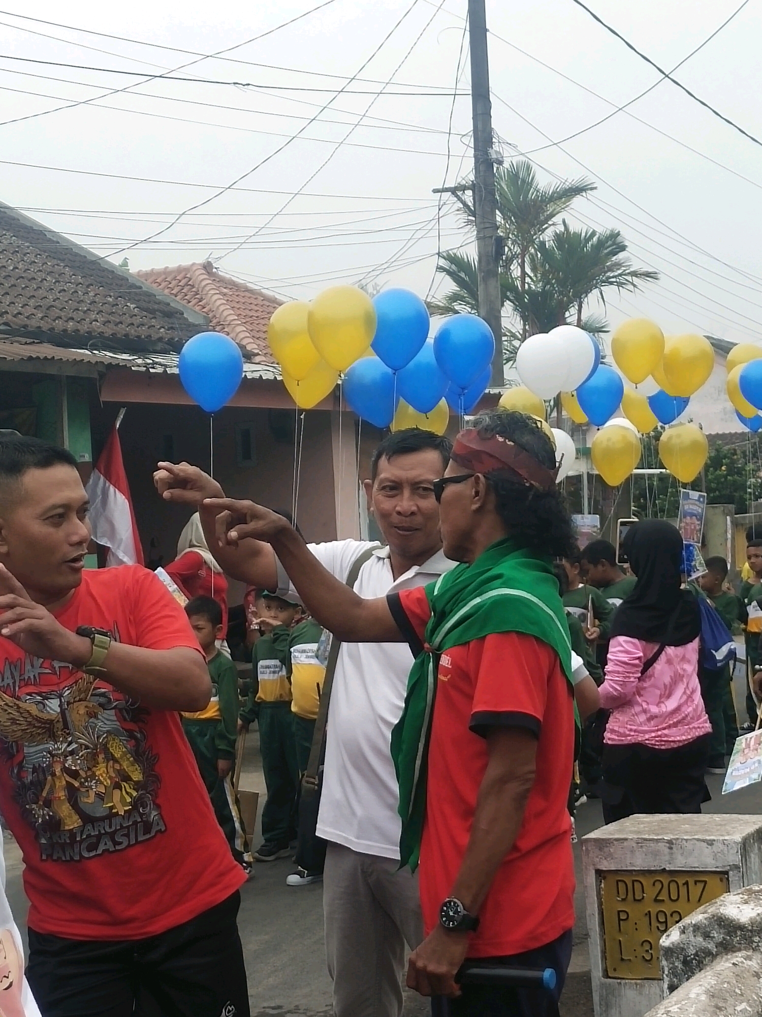 In Ambulu, Indonesia, a group of people are gathered outdoors under a cloudy sky, with power lines and houses in the background. In the foreground, three men are the primary focus. The man on the left, wearing a red shirt with a Garuda Pancasila graphic, gestures with his left hand. The man in the center, wearing a white t-shirt, points with both hands. The man on the right, wearing a red shirt, a green scarf, and sunglasses, looks towards the center man. Several blue, yellow, and white balloons are visible floating above the group, attached to strings. Some children and other individuals are present in the background, along with a flag. A concrete marker with text and numbers is visible in the lower right corner. The overall scene appears to be a casual gathering or event.