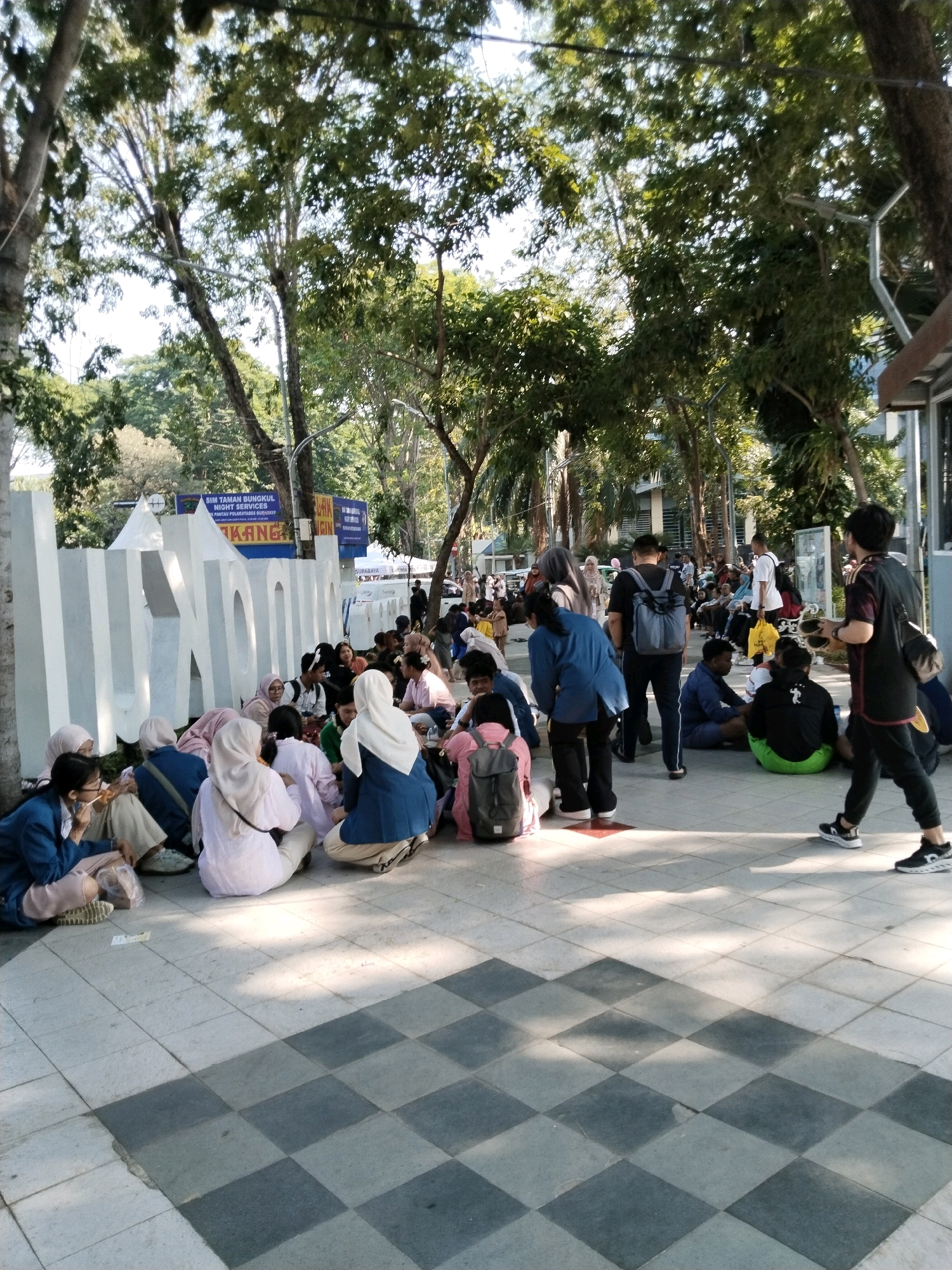 In Surabaya, Indonesia, a group of individuals, predominantly young adults, are gathered outdoors on a paved area with a checkered pattern, situated near a large white sculpture that appears to spell out a word. Many are seated on the ground, some in small groups, while others stand or walk. The individuals are dressed casually, with many wearing hijabs, indicating a cultural context common in Indonesia. Some are engaged in eating, while others appear to be conversing or resting. The environment is characterized by lush trees providing shade, suggesting a park or outdoor public space within the city. In the background, there are buildings and signs, including one that appears to be informational or directional, further establishing an urban setting. The lighting suggests it is daytime, possibly late afternoon, with sunlight filtering through the trees.
