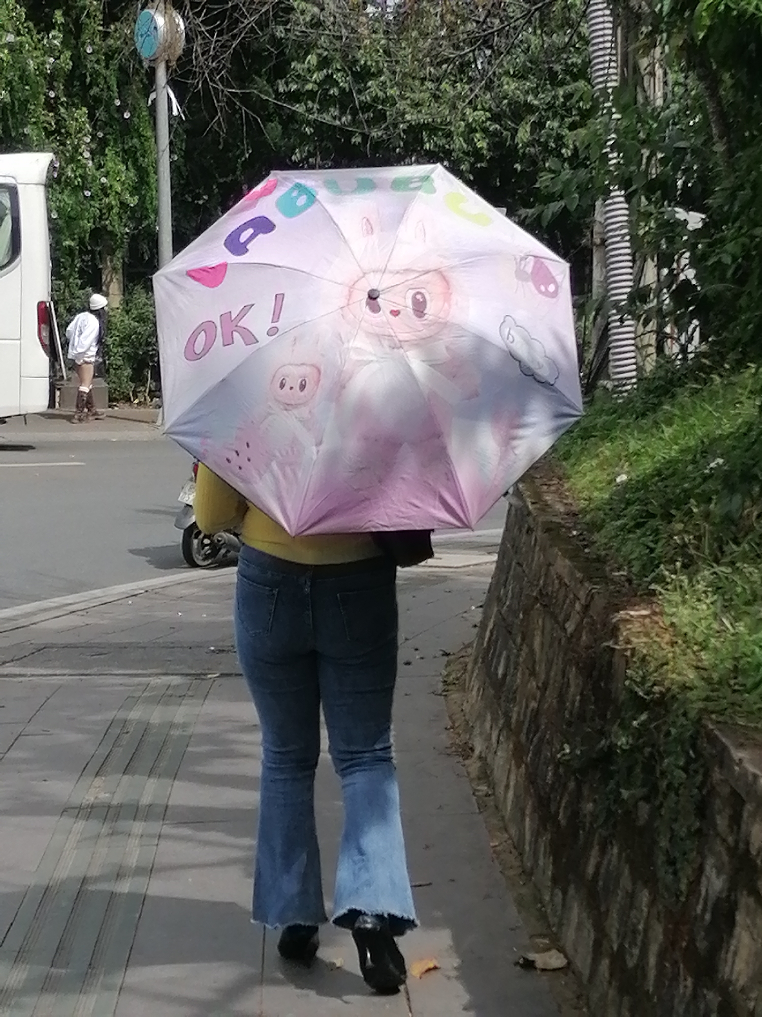 The image depicts a rear view of a person walking along a sidewalk in Da Lat, Vietnam, holding a large, colorful umbrella. The umbrella features cartoonish characters and the words "OK!" printed in pink. The person is wearing a yellow top and blue flared jeans. A white van is visible to the left of the frame, and a scooter is parked behind the person. The sidewalk is paved with tiles, and a stone wall covered in greenery lines the right side of the path. The background consists of trees and vegetation, suggesting an outdoor urban setting. A clock on a pole is visible in the upper left corner.