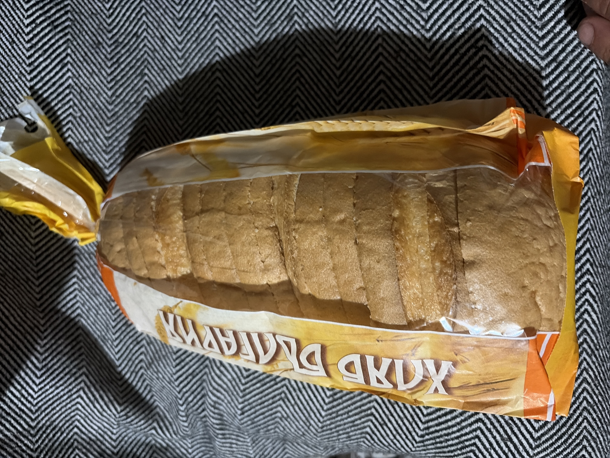 The image displays a loaf of sliced bread, encased in a clear plastic bag with an illustrative paper wrapper, resting on a black and white zigzag patterned fabric. The bread is light brown and appears to be thinly sliced. The packaging features a prominent yellow and orange design, with white text printed along its side. When viewed correctly, the Cyrillic text on the wrapper reads "ХЛЯБ ДЕЛИКАТЕС", which translates from Bulgarian to "Delicacy Bread". The bread is still sealed in its packaging, with one end gathered and secured by a yellow plastic clip or twist-tie. Shadows are cast by the bread and packaging onto the fabric, suggesting indoor lighting conditions. No people are visible, and no specific activity is taking place; it is a still life. The scene offers a close-up perspective of the product, likely in a casual household setting in Sirakovo, Bulgaria.