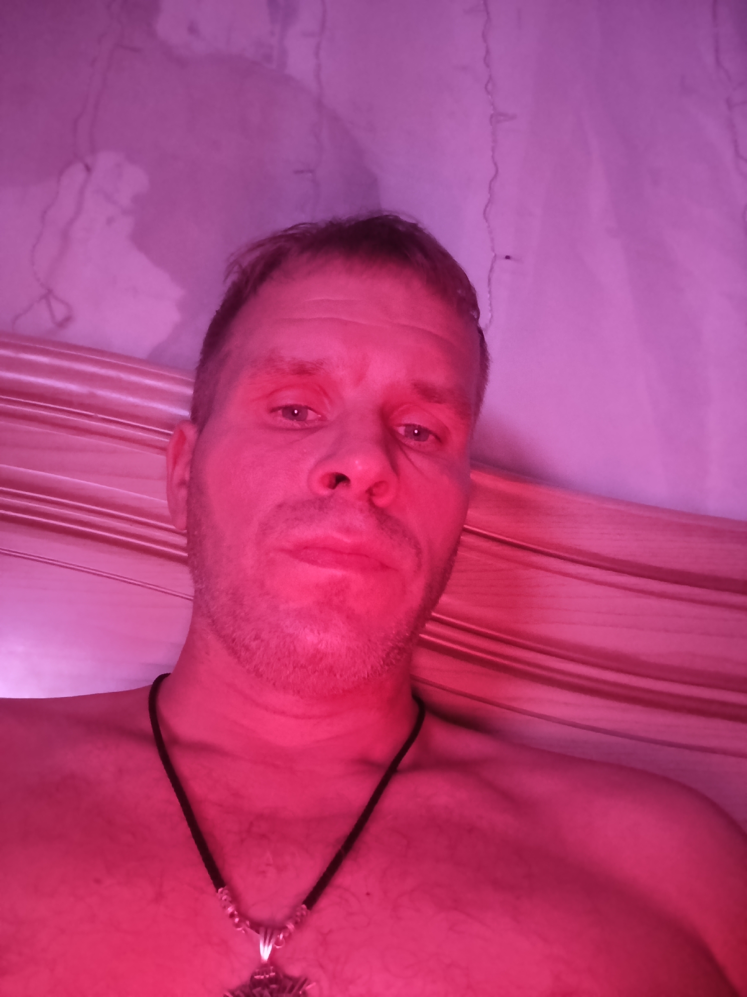 A close-up, low-angle, indoor selfie shows a shirtless, fair-skinned man with short, light brown hair and stubble. He is looking directly at the camera with a neutral expression. He is wearing a black cord necklace with a silver pendant. The man is positioned in front of a wooden slatted surface and a pinkish, textured background. The lighting is a strong pink hue, casting shadows and highlighting the contours of the man's face and upper body. The image appears to have been taken in the city of Unknown, Unknown.