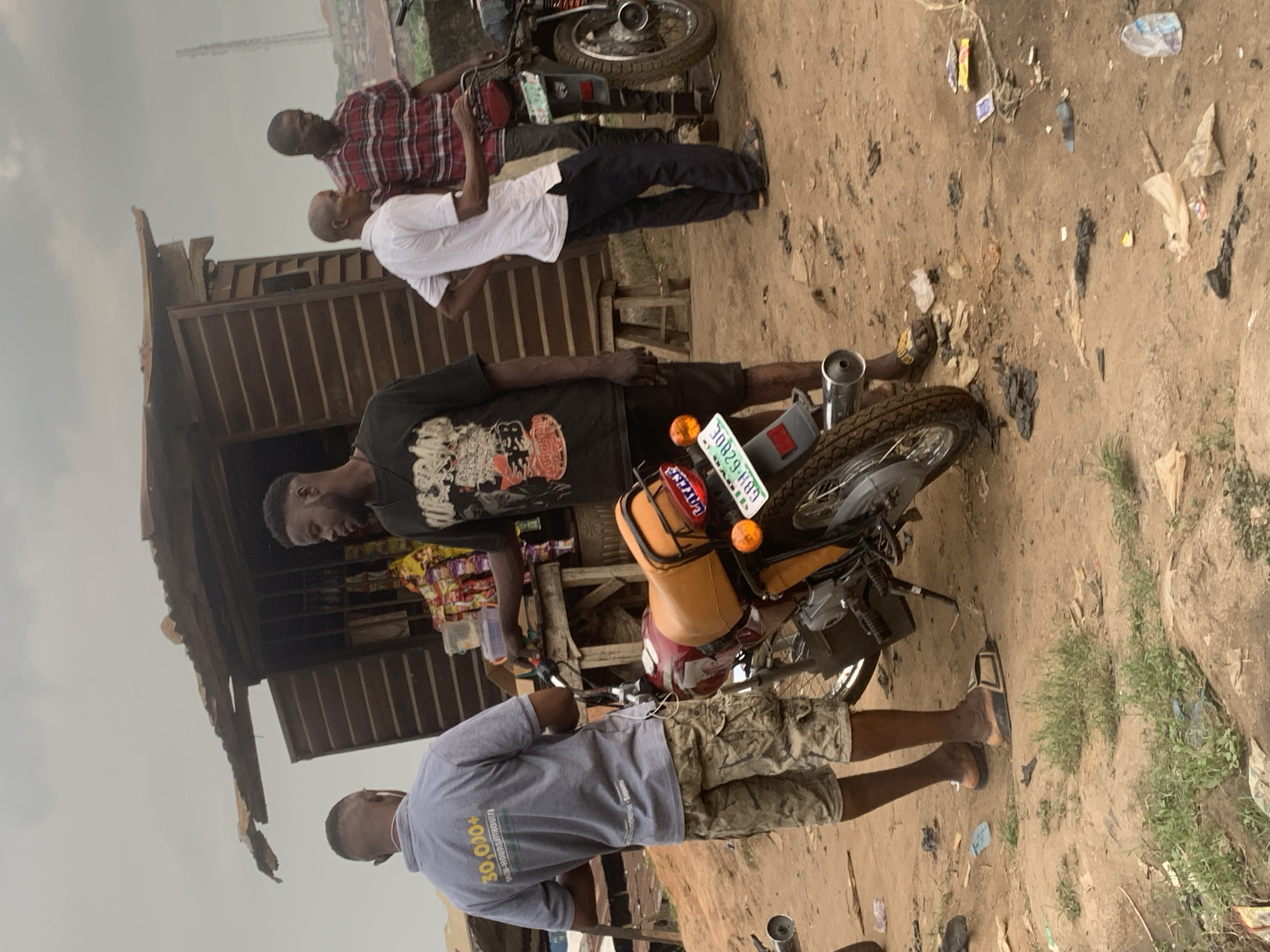 Four men are gathered in an outdoor setting, likely a roadside stall, in the city of Shagamu, Nigeria. The scene appears to be an informal marketplace or a vendor's spot. The ground is dirt, with scattered debris and some sparse vegetation.
In the foreground, a man wearing a grey polo shirt with "30,000+ MIN-HOURS/THOUBT!" printed on the back and camouflage-patterned shorts stands with his back mostly to the camera. He is wearing sandals. Beside him is a red and orange motorcycle, which is the primary focus of the middle ground. The motorcycle has a license plate with the characters "LGH-6280E".
To the right of the motorcycle, a man in a black t-shirt with a graphic design is engaged in conversation or transaction with another man. This second man is wearing a white short-sleeved shirt and dark trousers, and he is also wearing sandals. He is standing with his hands on his hips.
Further to the right, a third man is visible, wearing a red and black plaid shirt and dark trousers. He is looking down towards the motorcycle.
Behind the men, a small, weathered wooden structure with a corrugated metal roof serves as a stall. Shelves inside the stall are stocked with various packaged goods, including snacks and drinks. A wooden stool is placed near the stall. The background shows a slightly hazy sky and more distant urban structures, including what appears to be a cell tower. The overall impression is one of a bustling, everyday scene in a Nigerian urban environment.