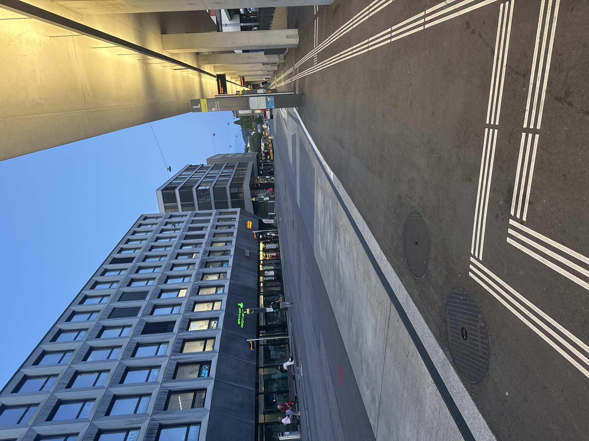 The image depicts a modern urban street scene in Liestal, Switzerland, likely at dusk or early evening, given the clear blue sky and prominent artificial lighting. The perspective is from underneath a large, illuminated concrete overhead structure, possibly an elevated walkway or a canopy, which casts a warm glow on its underside.
To the left, a tall, multi-story building with a distinctive, dark grey patterned facade and numerous rectangular windows dominates the scene. Some windows reflect the environment, while others show interior lights. At ground level of this building, several shops are visible. Text seen on the building includes "baloise" and "VISTA" on some windows, and shop signs like "BLKB" and "BAHNHOF APOTHEKE," which features a green cross logo.
To the right, an asphalt road features precise white markings, including sets of triple parallel lines and solid and dashed white lines, suggesting designated lanes or pedestrian areas. Two circular manhole covers are embedded in the road. Adjacent to the road is a concrete sidewalk or platform.
In the mid-ground, a tall, grey information pillar or signpost displays a large number "1" and text, partially legible as "Liestal" and possibly "Bahnhof" or "Busbahnhof." A digital display sign overhead shows "Bus nach Basel SBB," indicating a bus route. A yellow bus can be seen further down the street, and distant hills and buildings are visible against the twilight sky.
Several people are present, engaged in typical urban activities; some are sitting on benches near the shops, while others are standing or walking, suggesting a public transit stop or a busy commercial area. The overall impression is one of clean, well-maintained modern infrastructure.