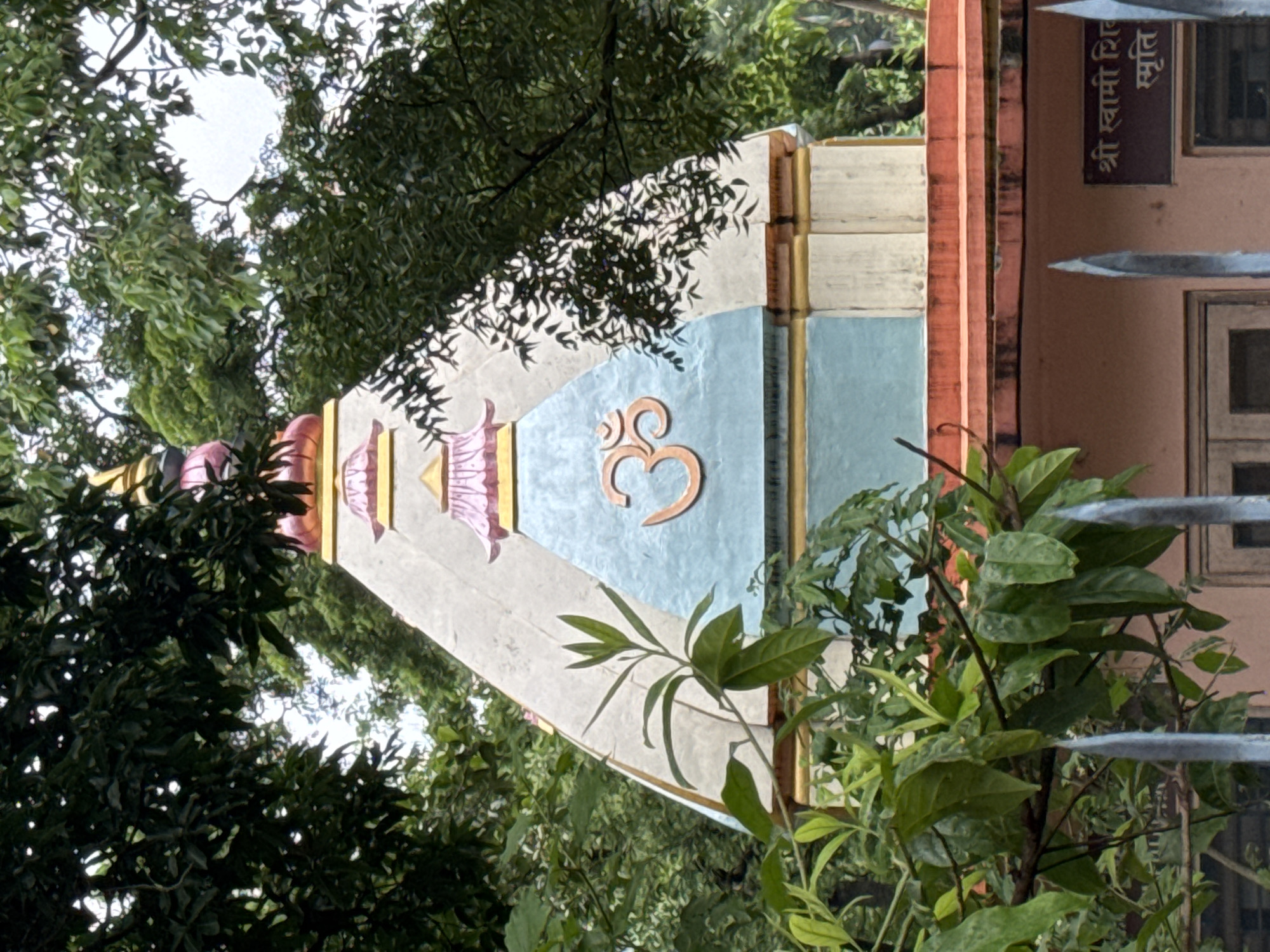 The image shows the upper portion of a temple structure in Rishikesh, India, partially obscured by lush green foliage. The temple has a multi-tiered pyramidal roof, adorned with decorative elements in shades of white, light blue, pink, and gold. Prominently displayed on the light blue section of the roof is the Om symbol, rendered in an orange hue. The roof is topped with a golden finial. Below the roof, an orange-colored cornice runs along the edge of the building. A sign in Devanagari script is visible on a brown plaque affixed to the building's wall, partially obscured by leaves. The foreground features sharp, metallic spikes of a fence, also partially covered by the dense green leaves of trees and bushes. The overall impression is one of a serene, natural setting surrounding a religious structure.
