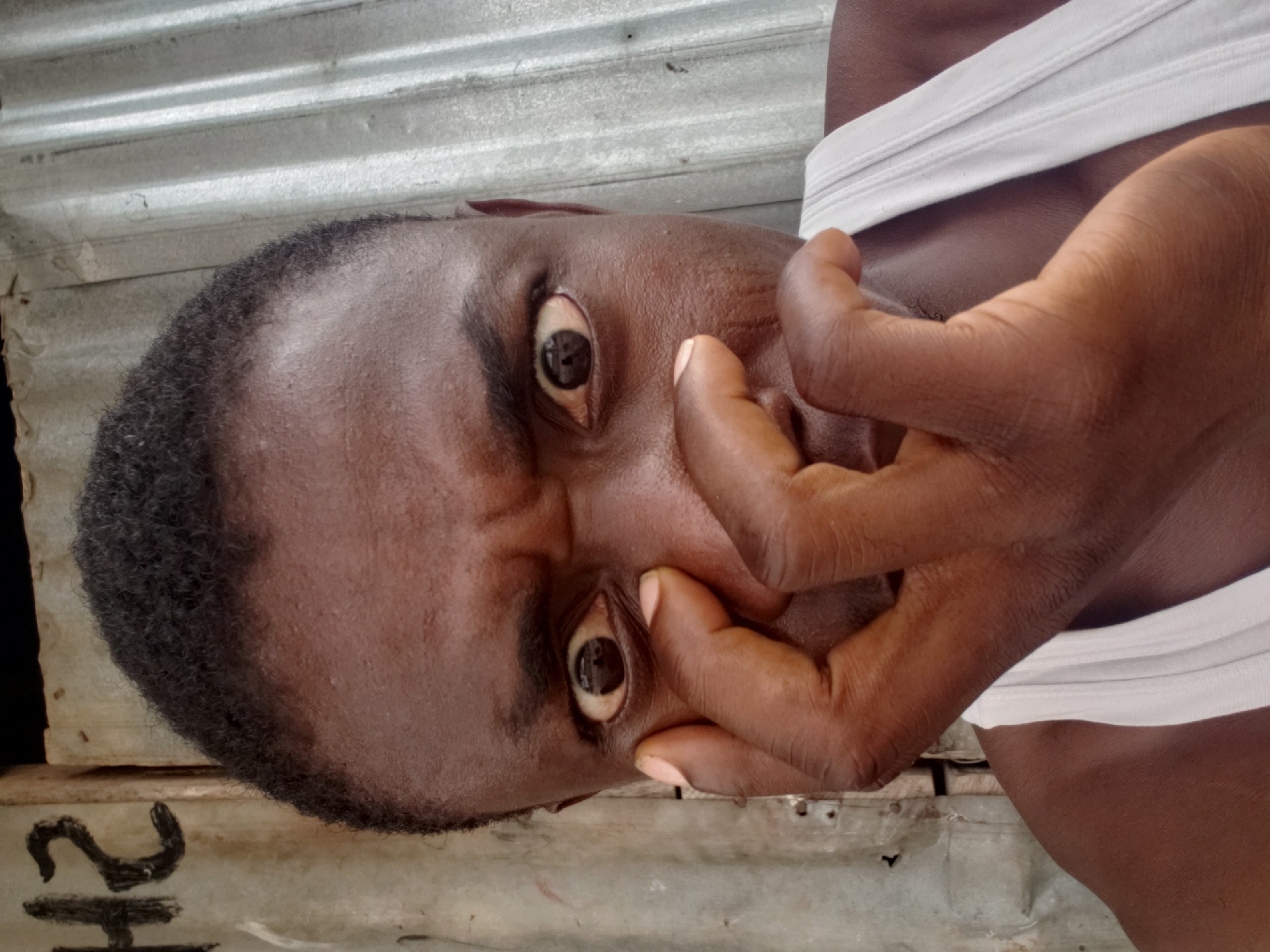 A close-up portrait, rotated 90 degrees clockwise, features an individual with dark skin and short, dark, curly hair. The person's face is centered in the frame, displaying an expression of intense surprise or shock, with both eyes wide open, revealing the whites and dark irises. Their hands are positioned around their lower face, with fingers partially obscuring their mouth and chin, emphasizing the dramatic reaction. The individual is wearing a simple white undershirt or tank top.
The background consists mainly of corrugated metal sheeting, possibly a wall or roof, along with some rough, light-colored wooden planks or surfaces. This utilitarian setting suggests an informal environment, aligning with the context of Adankolo, Nigeria. Natural, diffused light illuminates the scene, indicating it is daytime. A visible detail in the bottom left corner (when viewed upright) is a faint black marking resembling the number "21" on a light surface. The focus is tightly on the subject's expressive face, highlighting their features and emotional state against the textured, industrial background.