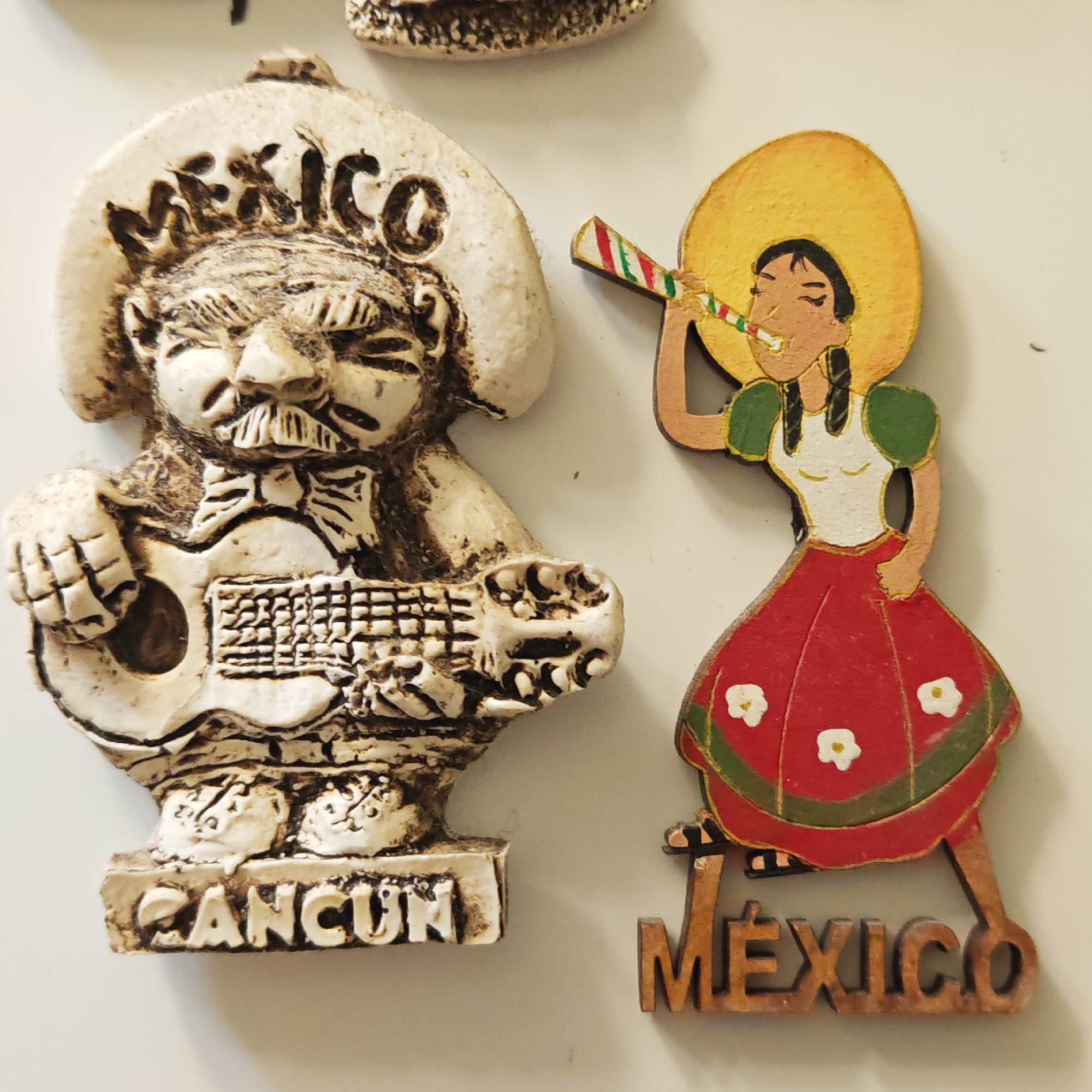 Two souvenir magnets depicting Mexican cultural figures are displayed on a plain, light-colored surface.
The magnet on the left is a textured, off-white sculpture of a man. He wears a large sombrero with the word "MÉXICO" embossed on the brim. He has a prominent mustache and appears to be playing a guitar. His attire includes a shirt with a bow tie. The base of this magnet features the word "CANCUN." The material resembles aged or antiqued ceramic or resin.
The magnet on the right is a colorful, painted wooden cutout of a woman. She wears a wide-brimmed yellow sombrero and has black braided pigtails. Her outfit consists of a white blouse with puffed green sleeves and a vibrant red skirt adorned with white flowers and green trim. She is shown blowing a party horn, which has red, green, and white stripes. The base of this magnet is a separate wooden cutout spelling out "MÉXICO" in capital letters.
Both magnets evoke a celebratory and festive atmosphere, representing Mexican culture and tourist destinations. The background is simple and unadorned, highlighting the magnets.