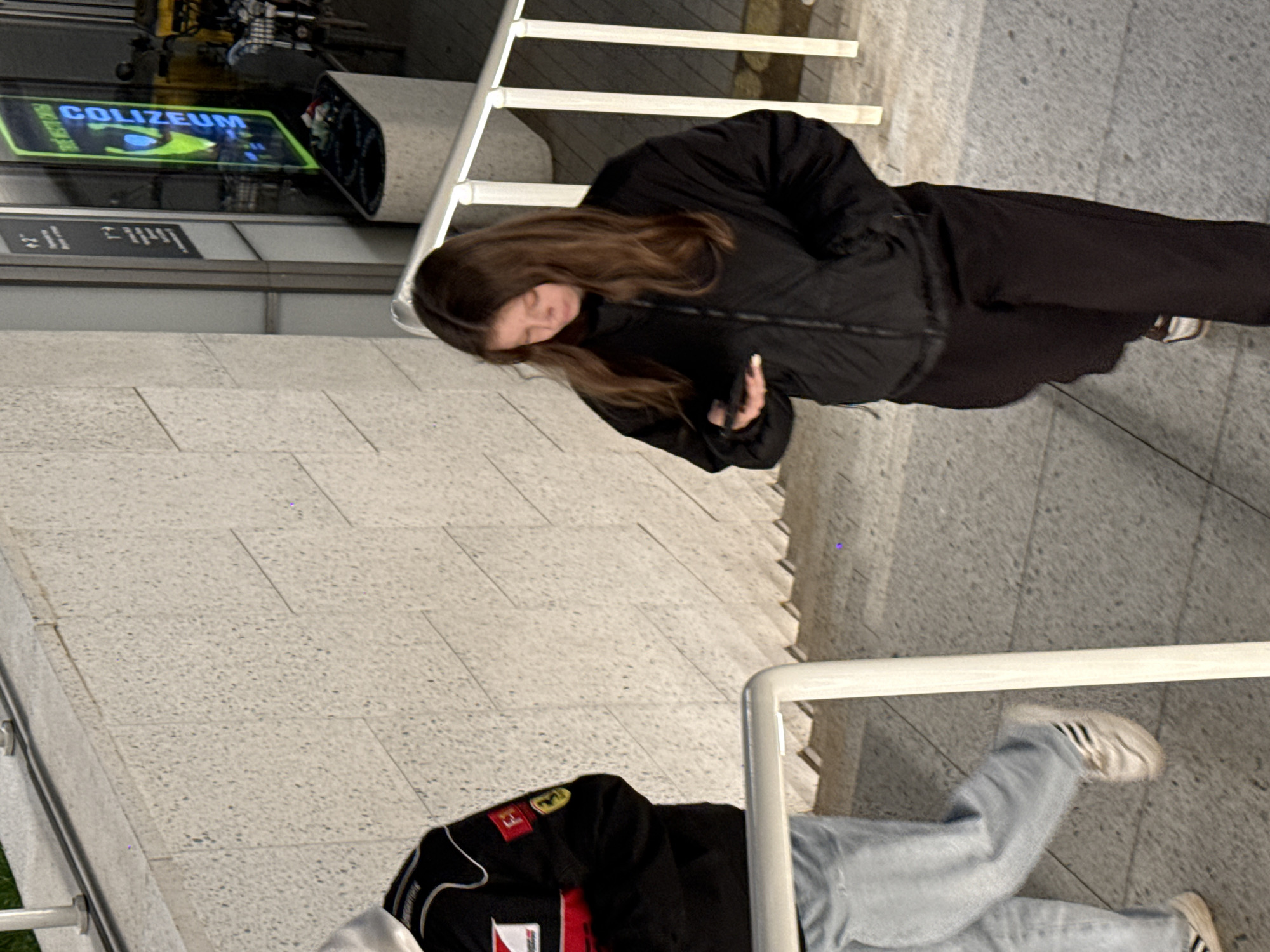The image captures an indoor or covered outdoor scene, likely a public thoroughfare or entrance area in Khimki, Russia, characterized by light-colored concrete flooring and white metal railings. The setting appears modern, illuminated by artificial light, possibly in the evening or at night, as indicated by a brightly glowing sign.
In the upper half of the frame, a woman is visible, engrossed in her smartphone. She has long brown hair and is dressed in a black puffer jacket and dark trousers. Her attention is solely directed downwards towards the device in her hands.
Partially visible in the lower foreground is another person, seen from the waist down and partially torso. This individual wears a black racing-style jacket featuring a prominent yellow Ferrari emblem and other logos, paired with light blue jeans and white sneakers with black stripes, characteristic of the Adidas brand. They appear to be standing or moving near the white railing.
A significant detail in the background is a glowing green and blue sign that clearly displays the text "COLIZEUM." This sign is reflected on a dark, reflective surface below it, suggesting a gaming club or esports arena, which aligns with the provided location context. The overall atmosphere is casual and urban.