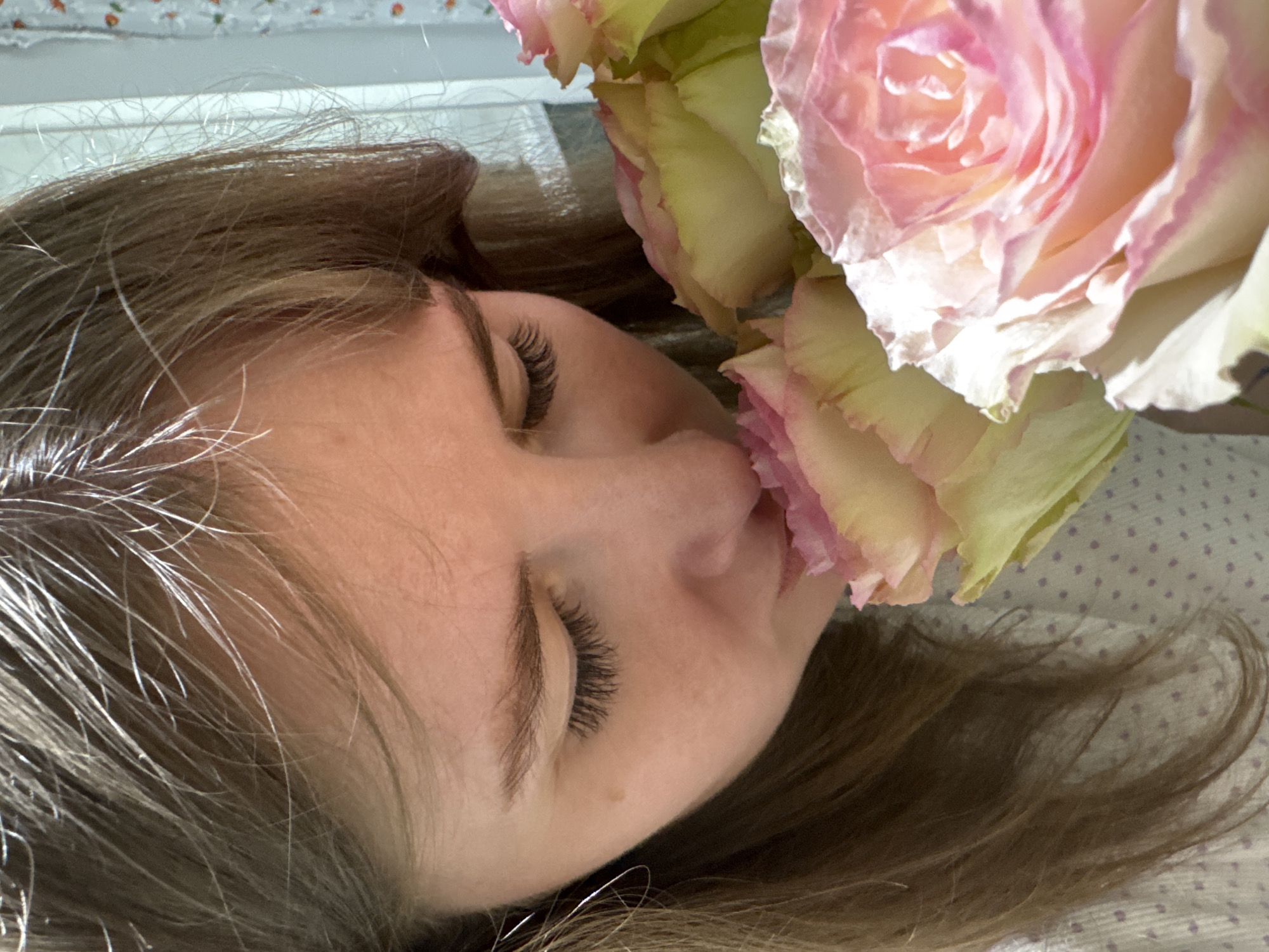 In an indoor setting in Gornyi (Dvurechenskoe S/P), Russia, a female subject is depicted from a high-angle, close-up perspective. Her head is tilted forward and to the right, with her nose pressed into a cluster of three-to-four roses, indicating the action of smelling them. The visible roses exhibit a color gradient from pale yellow-green at the base of the petals to a soft pink at the edges and center. The subject has light brown hair parted on the left side of her head, with a few lighter strands visible, particularly near the top. Her eyes are closed, with distinct, dark eyelashes visible. A small, dark mole is present on her left cheek, below the outer corner of her left eye. She wears a light-colored garment with a faint pattern of small, purple polka dots. The background, partially obscured, includes a white vertical architectural element and a patterned curtain featuring small, light-colored motifs against a light background.