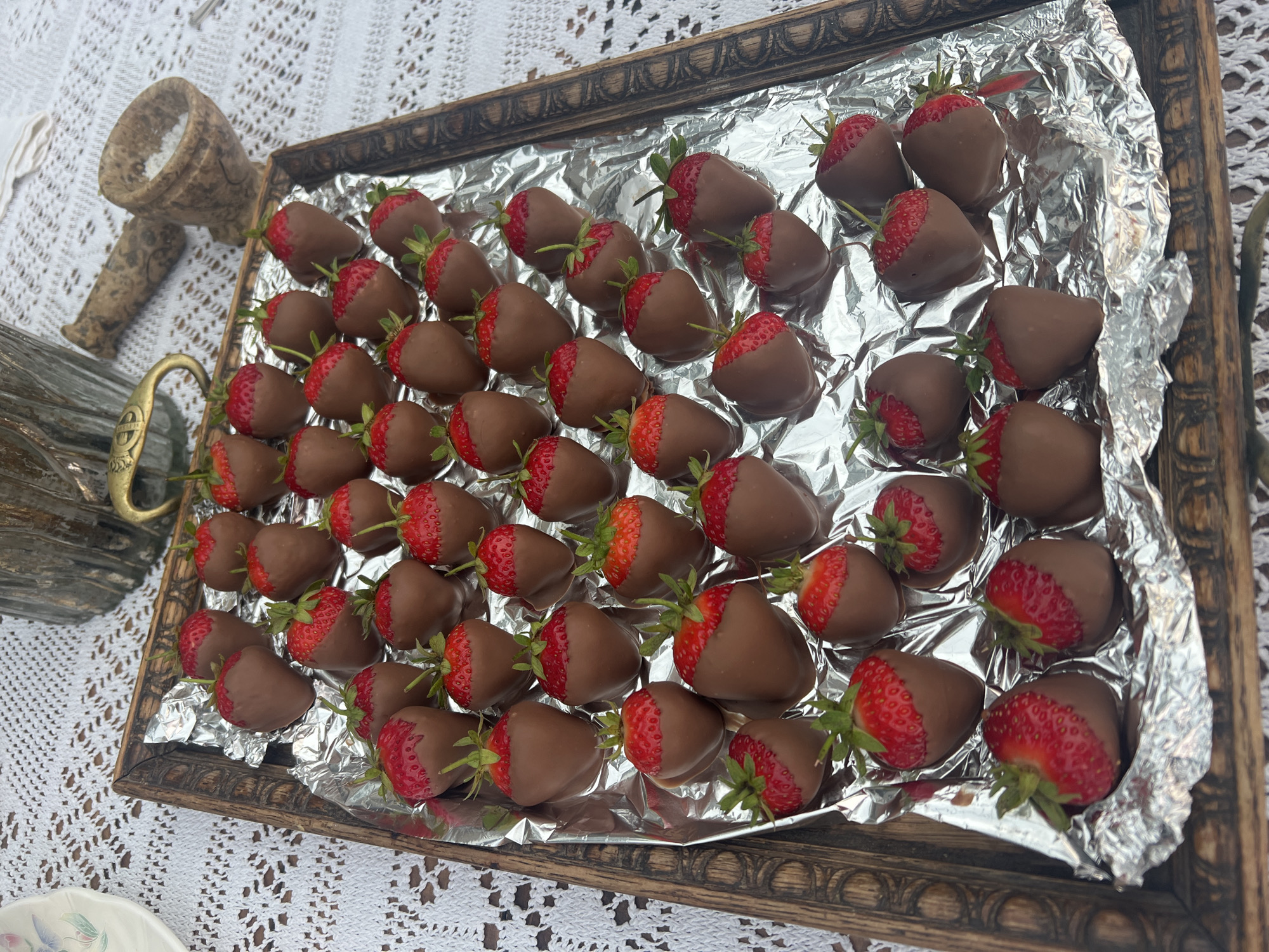 A wooden tray filled with chocolate-dipped strawberries is placed on a white lace tablecloth. The strawberries are arranged in rows, with some fully dipped in chocolate and others partially dipped, showing the red fruit and green stems. In the background, a decorative mortar and pestle and a vintage-style decorative item are visible. The scene is outdoors, with dappled sunlight suggesting it could be a garden or patio setting in Brighton, United Kingdom.