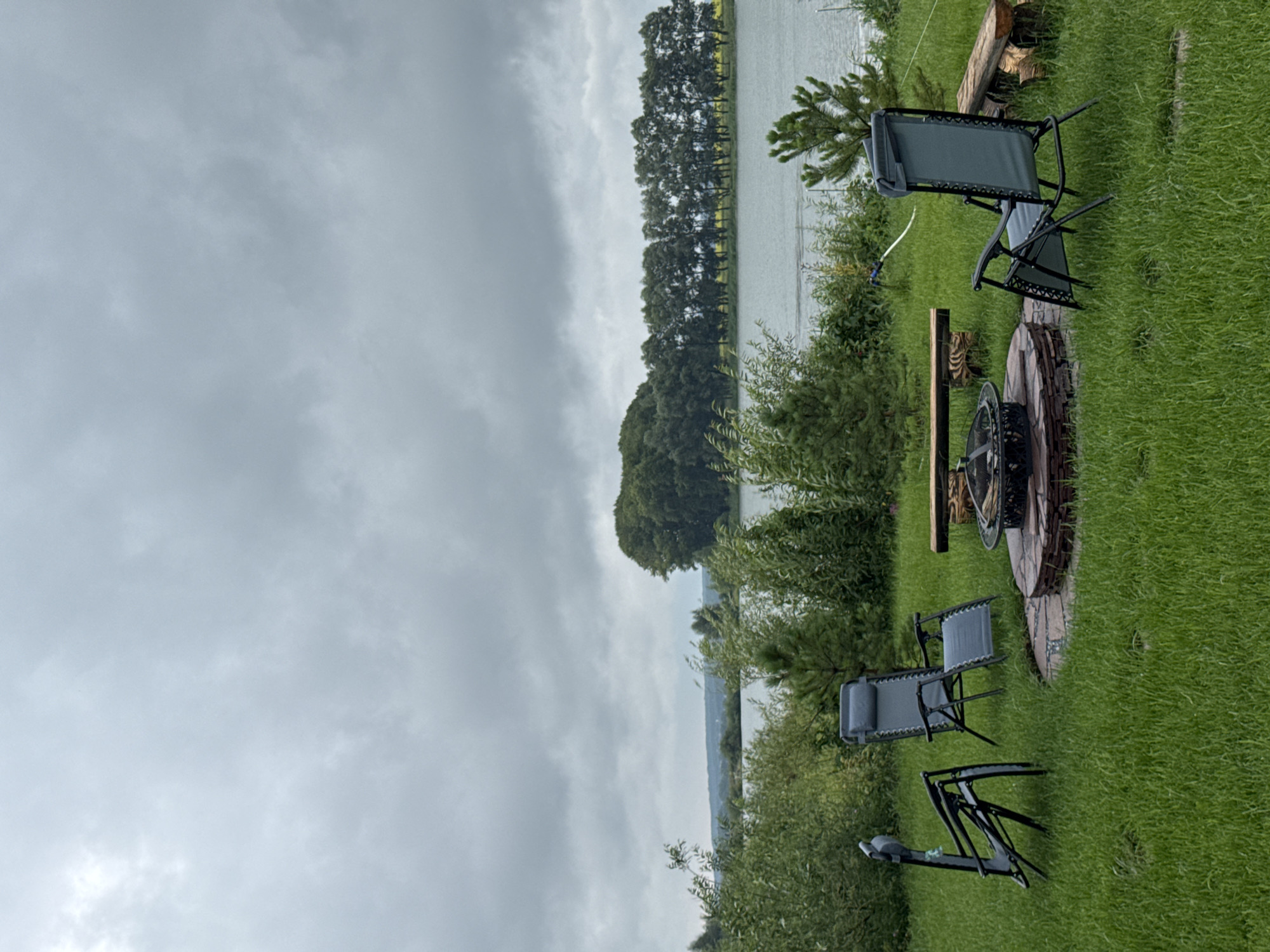 This is a vertical, eye-level, full shot of an outdoor seating area next to a body of water under a cloudy sky. In the foreground, a circular fire pit with stone surround and metal grate is centrally located. Four lawn chairs are positioned around the fire pit, two on the left and two on the right, all facing inward. One chair on the left is folded and leaning. Two rustic log benches are situated behind the fire pit. The area is covered in lush green grass. In the mid-ground, several small evergreen trees and leafy bushes are scattered throughout the lawn. Beyond the lawn, a calm body of water stretches across the frame. On the far side of the water, a dense line of trees forms a dark green border, with a small hill covered in trees to the left and a lighter, more distant treeline to the right. The sky is overcast with heavy, grey clouds. The scene appears to be in a rural or semi-rural setting.