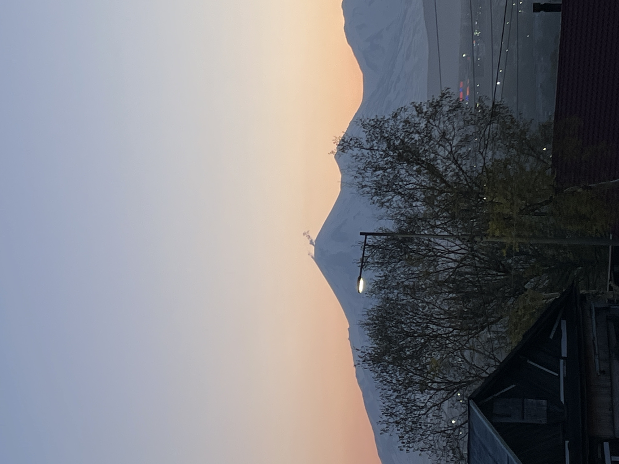 The image presents a wide outdoor view of Petropavlovsk-Kamchatsky, Russia, during twilight hours, characterized by a visible sky gradient and prominent snow-capped mountains. The sky transitions from a pale periwinkle blue at the top to a warm peach and light orange hue near the horizon. Centrally positioned in the midground is a large, snow-covered stratovolcano, likely Koryaksky or Avachinsky, from which two distinct plumes of white steam or gas emanate near its summit. Additional snow-clad mountain ranges extend to the left and right, receding into the distance.
In the lower-left foreground, the dark gable roof and upper section of a wooden structure are visible. Adjacent to this, a dense cluster of deciduous trees with mostly bare branches occupies the lower-center, partially obscuring the base of the mountains. An illuminated street light, mounted on a dark pole, is prominently situated within this tree cluster, positioned in front of the central volcano. On the lower-right, a portion of a dark reddish-brown tiled roof is visible. Scattered artificial lights, indicative of urban infrastructure within Petropavlovsk-Kamchatsky, are faintly discernible through the trees and in the distance above the lower-right roof. No human subjects are present in the scene.