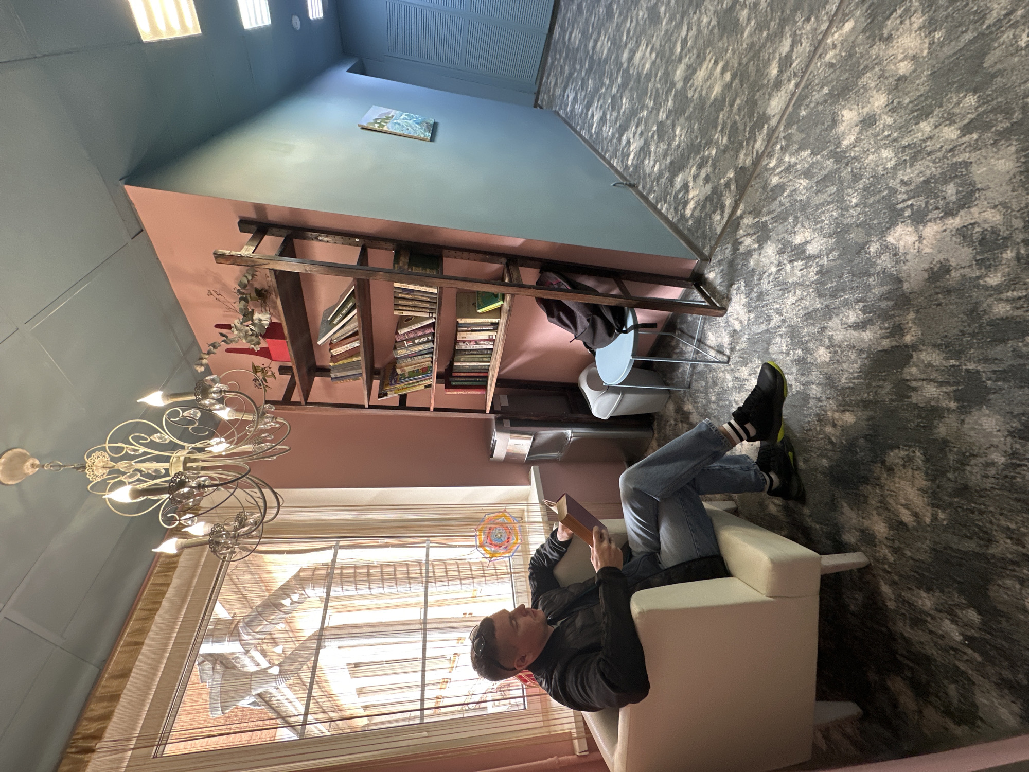 A man sits in a white armchair reading a book in what appears to be a room in Saint Petersburg, Russia. The room has a unique color scheme, with the upper walls painted a light blue and the lower walls a muted pink. A bookshelf filled with books stands against the pink wall. A chandelier hangs near a window with blinds. The floor is covered with a patterned carpet. The man is wearing jeans, a black jacket, and black and green sneakers.