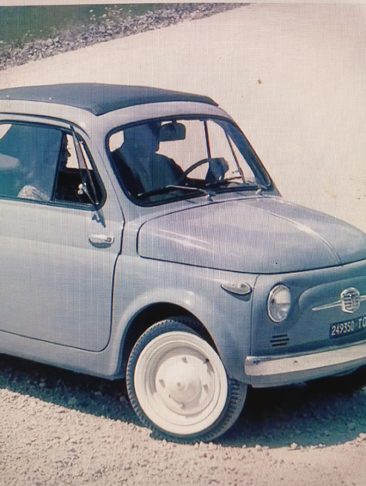 The image shows a light blue Fiat 500 car parked on a gravel road. The car has a sunroof and a rear-view mirror visible. A person is seated in the driver's seat, and their silhouette is visible through the window. The license plate of the car reads "249350 TO". The background consists of a dusty or gravelly road and a hint of greenery and rocks. There are no specific details in the image that indicate the location is the city of Sluck, Belarus.