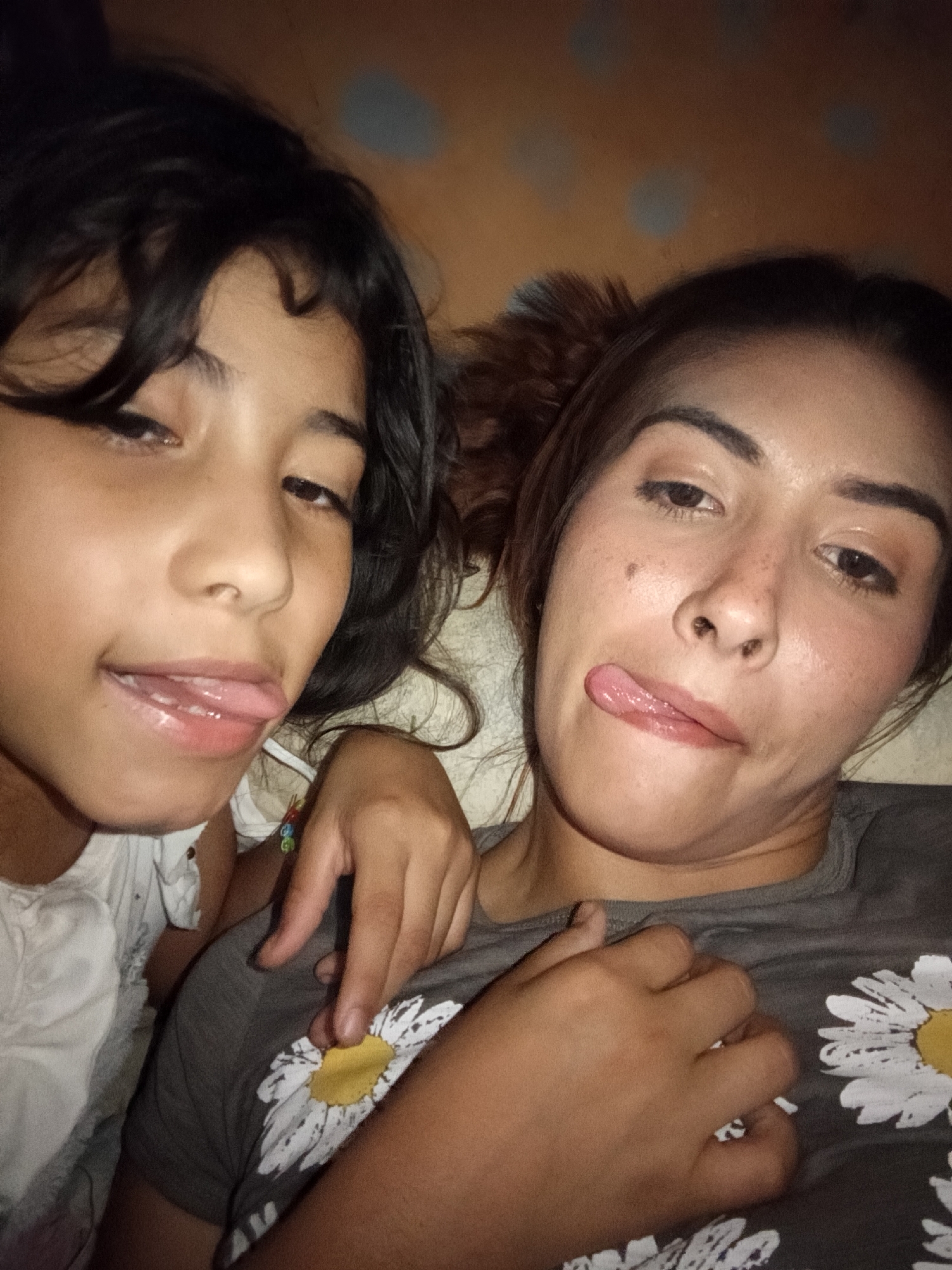 Two individuals, presumably a child and an adult female, are positioned close together, possibly lying down. Both are sticking their tongues out. The child has dark hair and is wearing a white garment. The adult female has brown hair pulled back and is wearing a gray shirt with daisy patterns. The background appears to be an interior setting with mottled walls. It is not possible to confirm that this image was taken in Juan Griego, Venezuela, without further information.