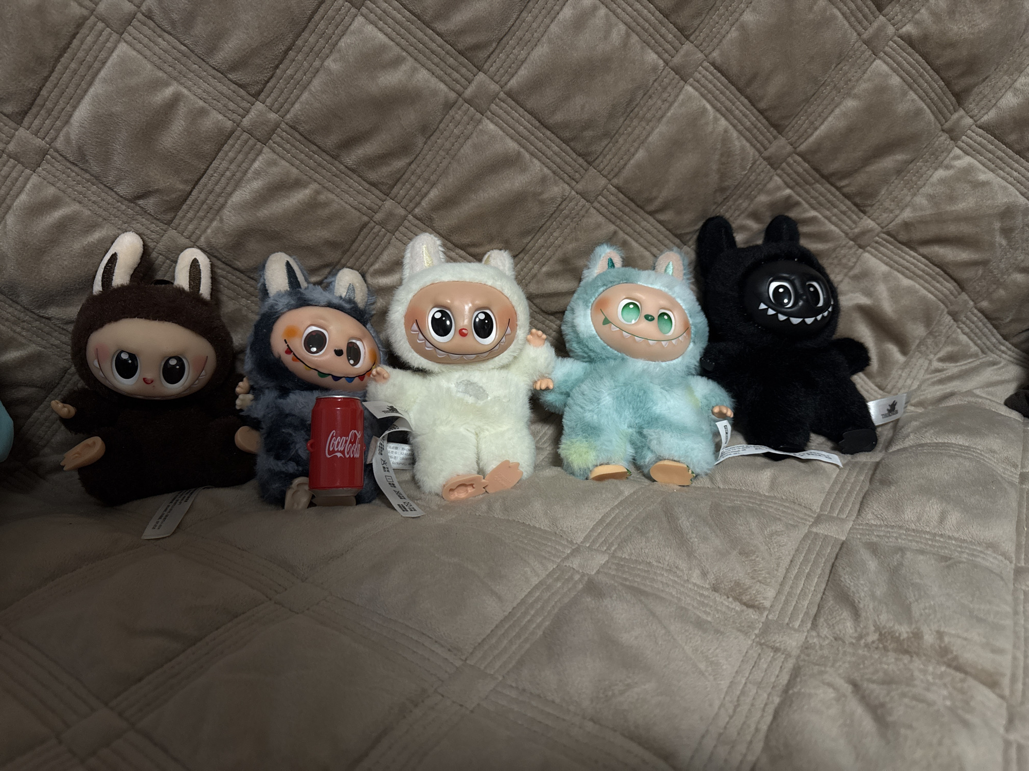 Five plush toys are arranged in a horizontal line on a quilted, light brown plush fabric surface, likely a blanket or sofa. From left to right, the first toy is dark brown with rabbit-like ears and a white face featuring large black eyes and a small mouth. The second toy is grey-blue with similar ears, a white face, large black eyes, and visible multicolored teeth, holding a miniature red Coca-Cola can between its feet. The third toy is white with small ears, a white face, large black eyes, and white teeth. The fourth toy is light blue-green with small ears, green eyes, and white teeth. The fifth toy, on the far right, is entirely black with small ears, a black face, large white eyes, and jagged white teeth. Several white product tags are visible beneath the toys. The setting is an indoor environment. The specific location within the city of Chaikovsky, Russia, cannot be determined from the visual evidence.