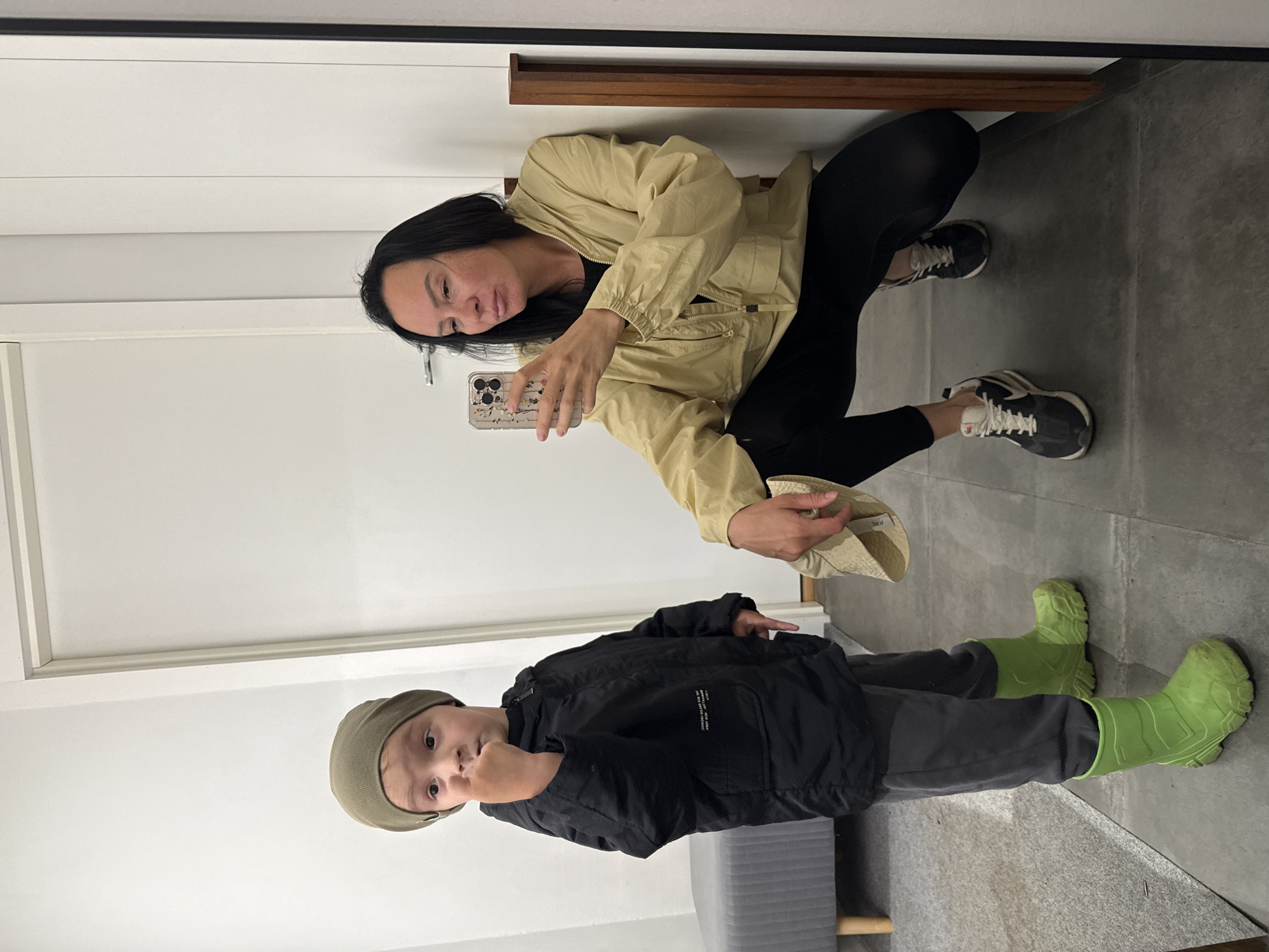 In an indoor setting, a child wearing a green beanie, a black jacket, gray pants, and bright green rain boots stands on the left. The child is holding a finger to their mouth. To the right, a woman wearing a light yellow jacket and black leggings squats down, holding a smartphone and taking a selfie. She has dark hair and is looking at the phone. The background consists of a white door, a gray upholstered bench, and a tiled floor. The scene appears to be a dressing room or fitting area, possibly in a store or home. The location is not identifiable as Chaikovsky, Russia.