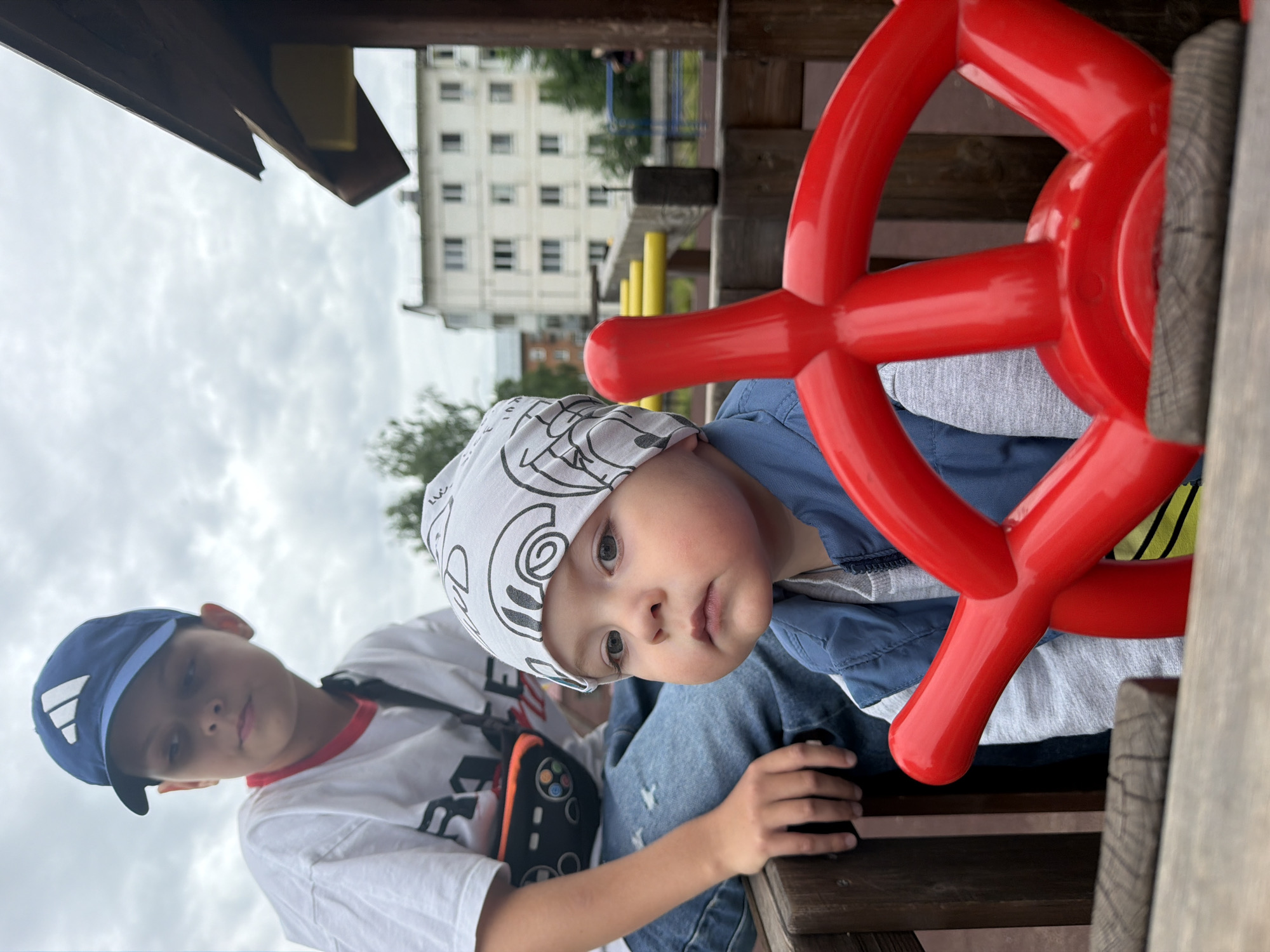 The image depicts two young boys on a playground with a building in the background. The foreground features a red steering wheel of a playground apparatus. On the left, an older boy wearing a blue baseball cap and a white t-shirt with a graphic print is seated, with a black fanny pack worn across his chest. To his right and slightly in front, a younger boy is visible, wearing a white bandana with a car print and a blue vest over a grey shirt. He is looking towards the camera with a neutral expression. In the background, a multi-story building with a light-colored facade and numerous windows is visible. The sky is overcast with clouds. The playground appears to be constructed of wood and metal. Based on metadata, the location is Votkinsk, Russia.