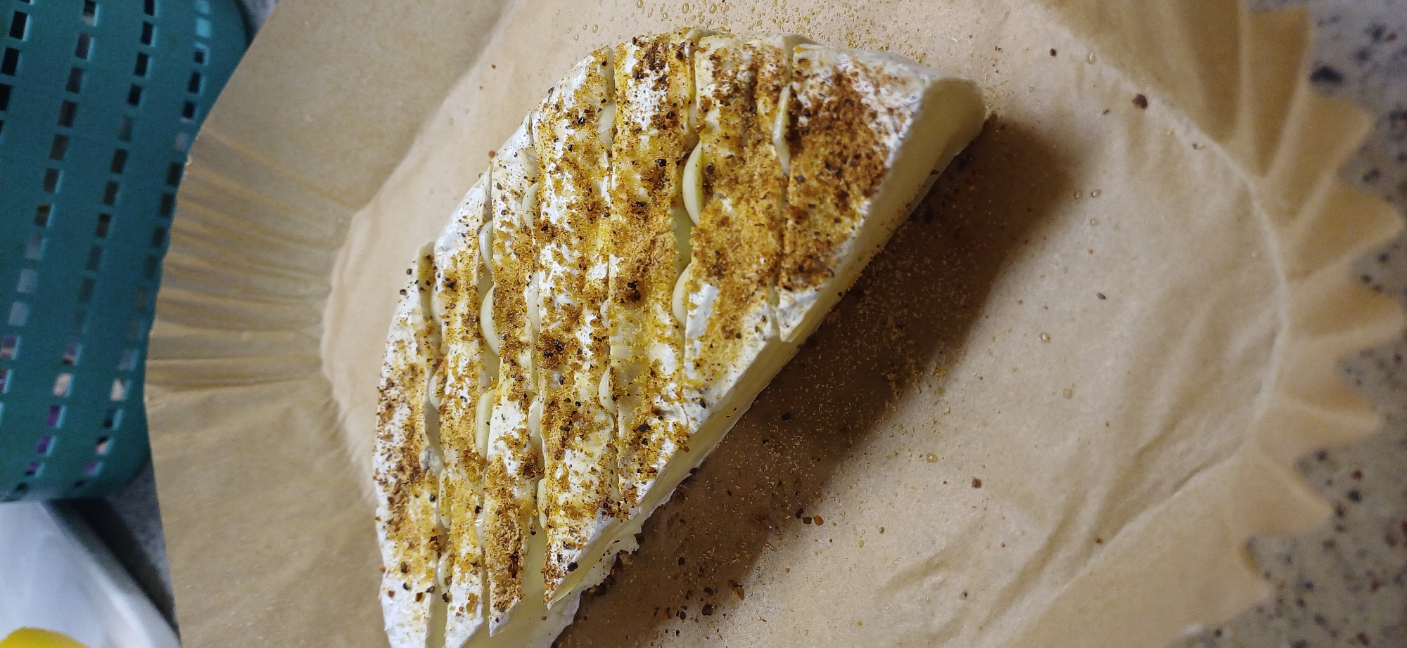 A wedge of brie cheese, seasoned with what appears to be dried herbs and spices, sits on a piece of parchment paper. The cheese is scored on top, revealing thin slices of garlic embedded within. The brie is triangular in shape, with a creamy white rind and a pale yellow interior. The parchment paper is light brown and has a slightly wrinkled texture. In the background, to the left, a teal-colored plastic basket with a grid pattern is visible. The lighting suggests it is daytime, but there are no specific cues to the weather or a precise location. The scene is simple and focused on the prepared cheese, implying it is either being prepped for cooking or has just been seasoned. There is no visible text in the image.