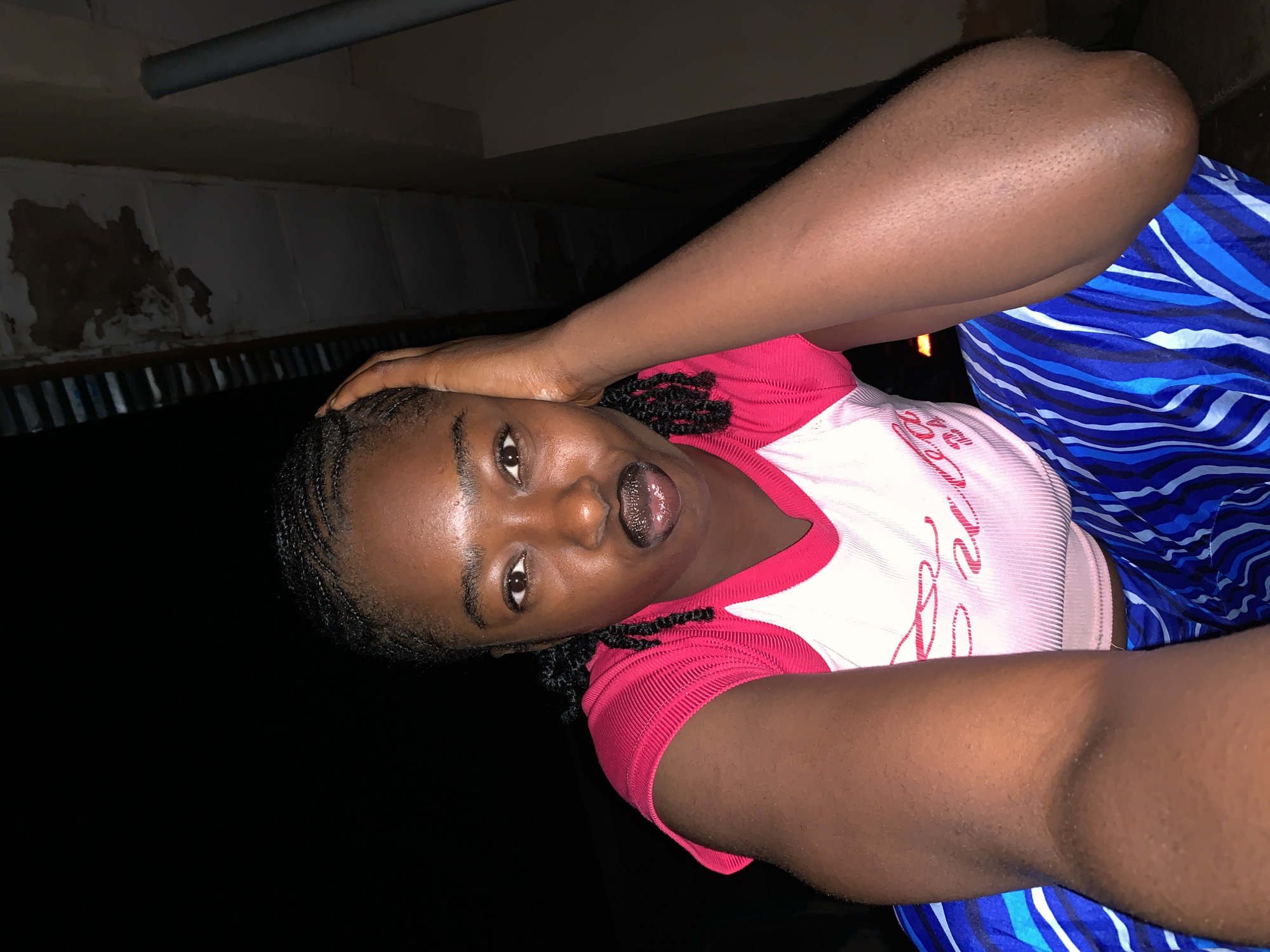 A young woman is captured in a close-up selfie, taken from a low angle. She has dark skin, braided hair, and is looking directly at the camera with a lively expression, her mouth slightly open, revealing her tongue. Her right hand is placed on the top of her head.
She is wearing a short-sleeved top with a white body and red trim around the neckline and sleeves. Stylized pink text, possibly the word "Love", is visible on the front of her shirt. Below her waist, she is wearing a garment with a distinct blue and white striped pattern.
The background is dark and appears to be an indoor or covered outdoor space, likely at night given the dim lighting. Above her, a gray pipe runs horizontally across the ceiling or overhang. Below this, there's a wall section, parts of which show discoloration or damage, and a lower structure that resembles corrugated metal or slats. The overall setting suggests a modest or utilitarian environment in Rafin Par, Nigeria. The lighting on her face suggests a direct light source, possibly a phone flash.