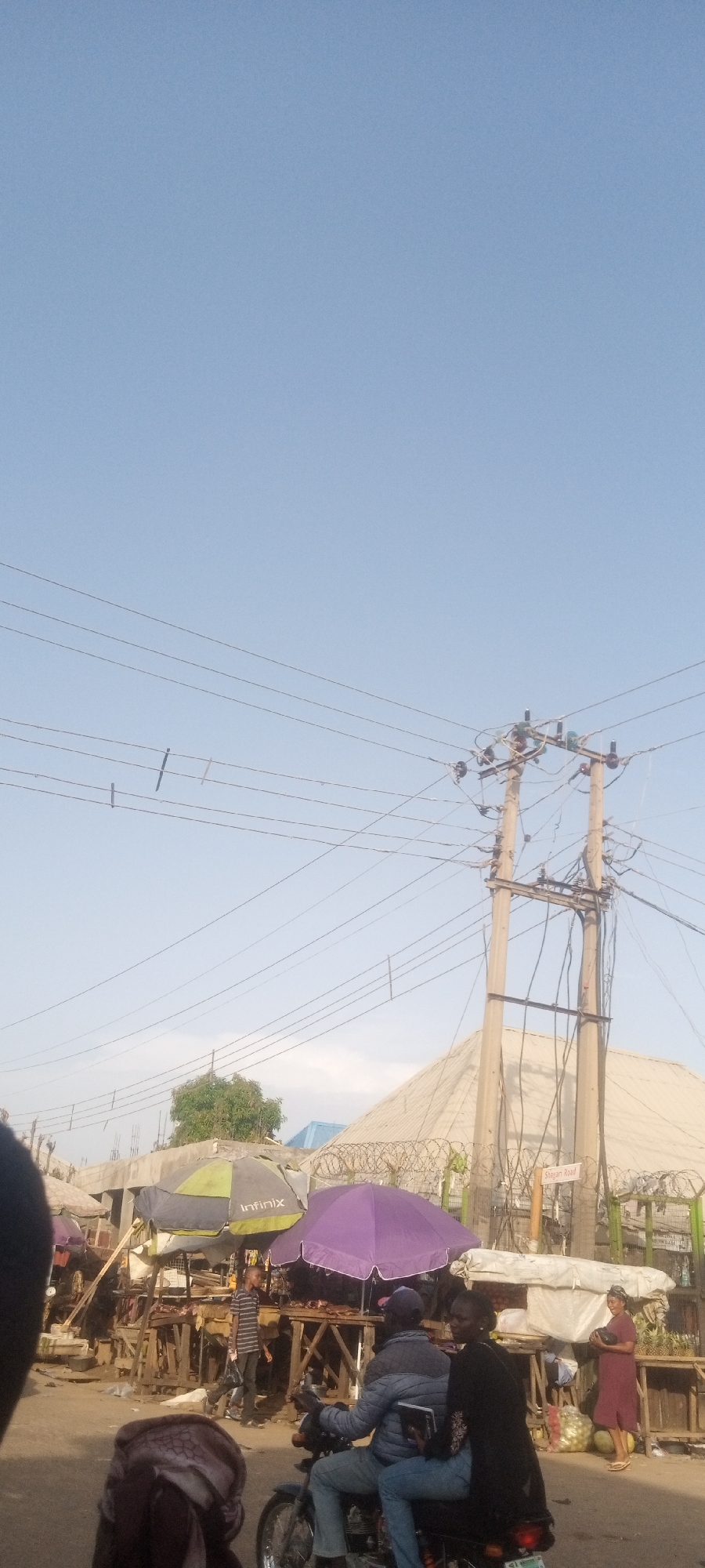 The image shows a street scene in Narayi, Nigeria. Overhead power lines stretch across a light blue sky. In the midground, a concrete electrical pole stands amidst market stalls. The stalls are covered with umbrellas, one is green and black with the word "INFINIX" on it and another is purple. People are visible near the stalls, including one wearing a red dress. A man and a woman ride on a motorcycle in the foreground. The background includes buildings with barbed wire on top of the walls, marked with "Shagari Road" sign.