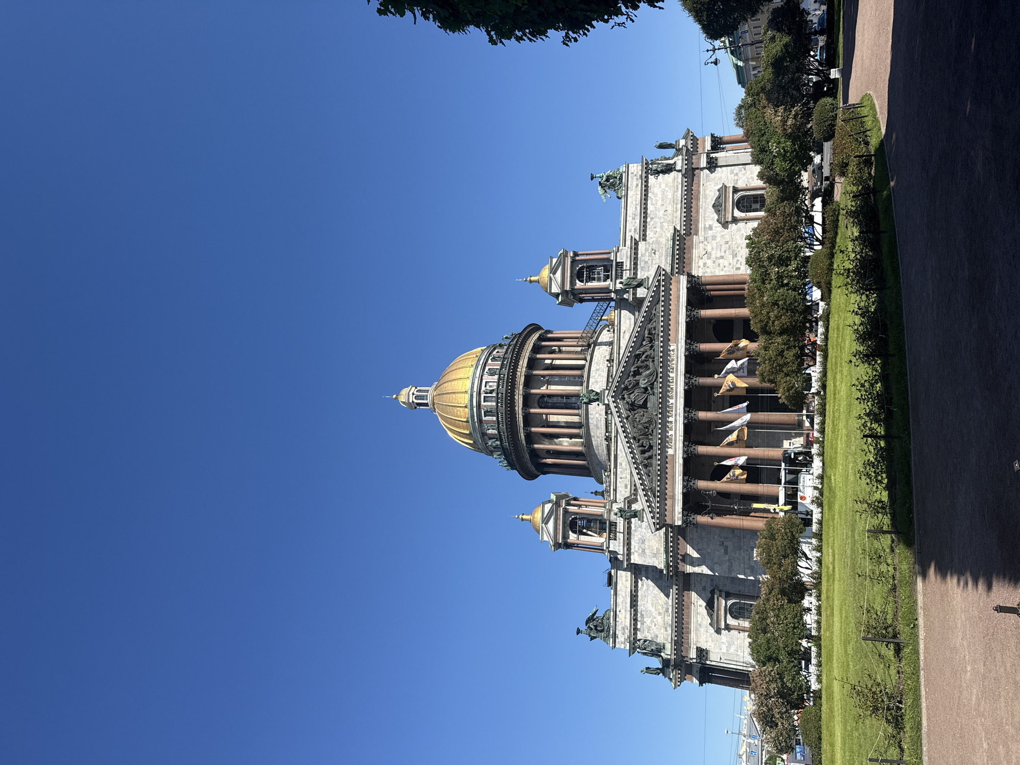The image captures a majestic view of Saint Isaac's Cathedral in Saint Petersburg, Russia, under a clear, bright blue sky. The grand cathedral dominates the scene with its impressive golden main dome and four smaller golden cupolas, gleaming in the sunlight. The building's facade is constructed from light-colored stone, adorned with numerous reddish-brown columns that support large porticos. Intricate sculptural details and classical statues can be seen along the roofline and pediments above the entrances.
The cathedral is set within a well-maintained public space, featuring expansive green lawns and several rows of neatly trimmed trees that line the perimeter of the building. A paved pathway runs along the right side of the frame, suggesting an area accessible to pedestrians. In the background, beyond the trees, other urban structures and a few vehicles, including what appears to be a white bus or truck, are partially visible.
No specific activity or event is taking place; the image is a static portrayal of the architectural landmark. The time of day appears to be mid-morning or early afternoon, indicated by the brilliant sunshine and the cloudless sky, suggesting fair weather. Several flags, predominantly yellow or gold with emblems, are displayed near the main entrance, adding to the ceremonial feel of the site. Latin text is visible on the frieze above the central portico, though not entirely legible due to distance.