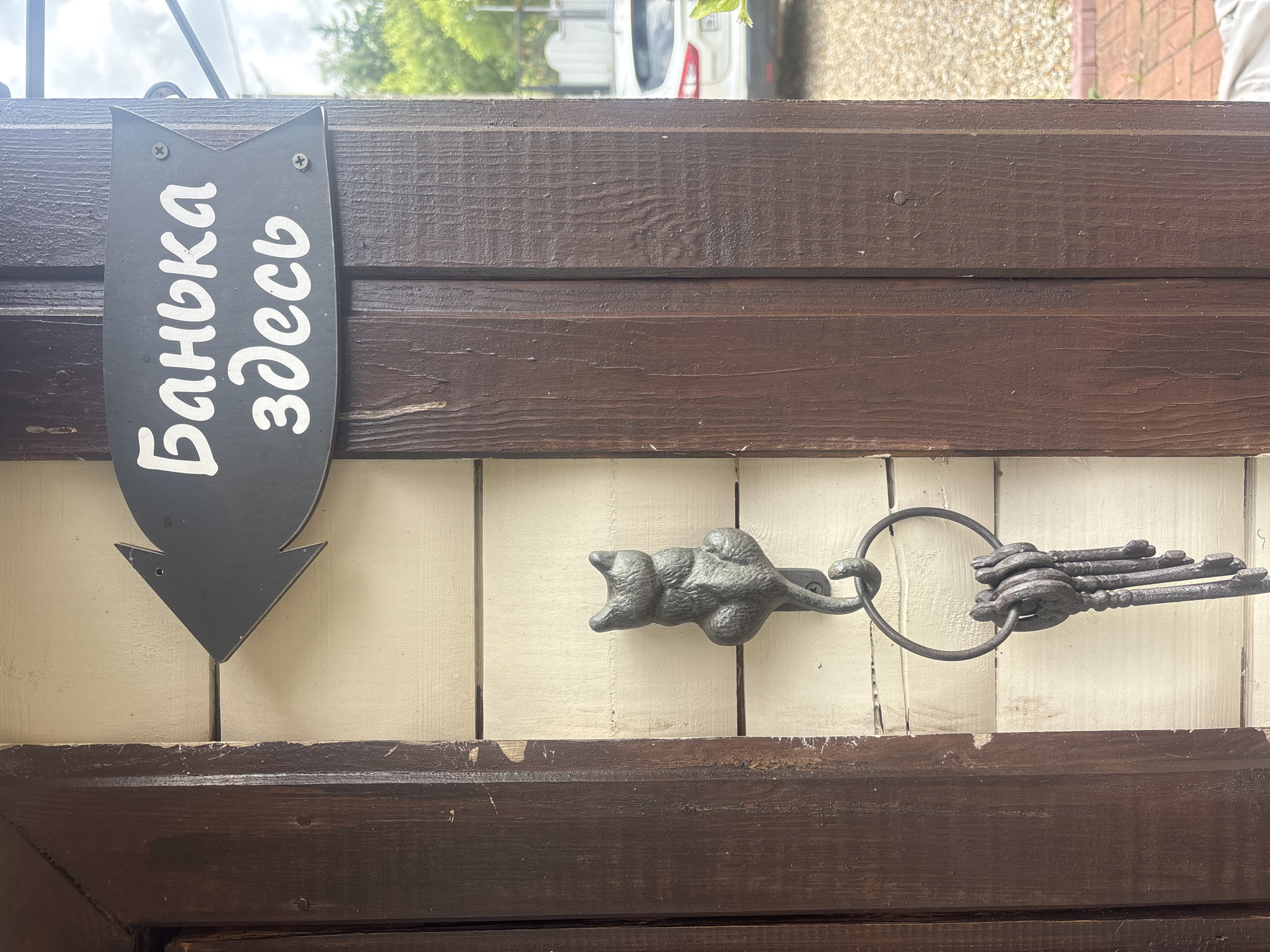 A black arrow-shaped sign is mounted to a wooden post and reads "Баня здесь" (Bathhouse here) in white cursive font, pointing to the left. Below the sign, a cast iron cat figurine is attached to the wall, serving as a hook. A ring with several old keys hangs from the cat's tail. The sign is on a light-colored, horizontal wooden plank wall, with dark brown wooden posts on either side. The background shows greenery, a white fence, and a portion of a white car parked on a gravel driveway. The scene appears to be outdoors, possibly in a rural or suburban setting in Лотос Снт (марьино), Russia.
