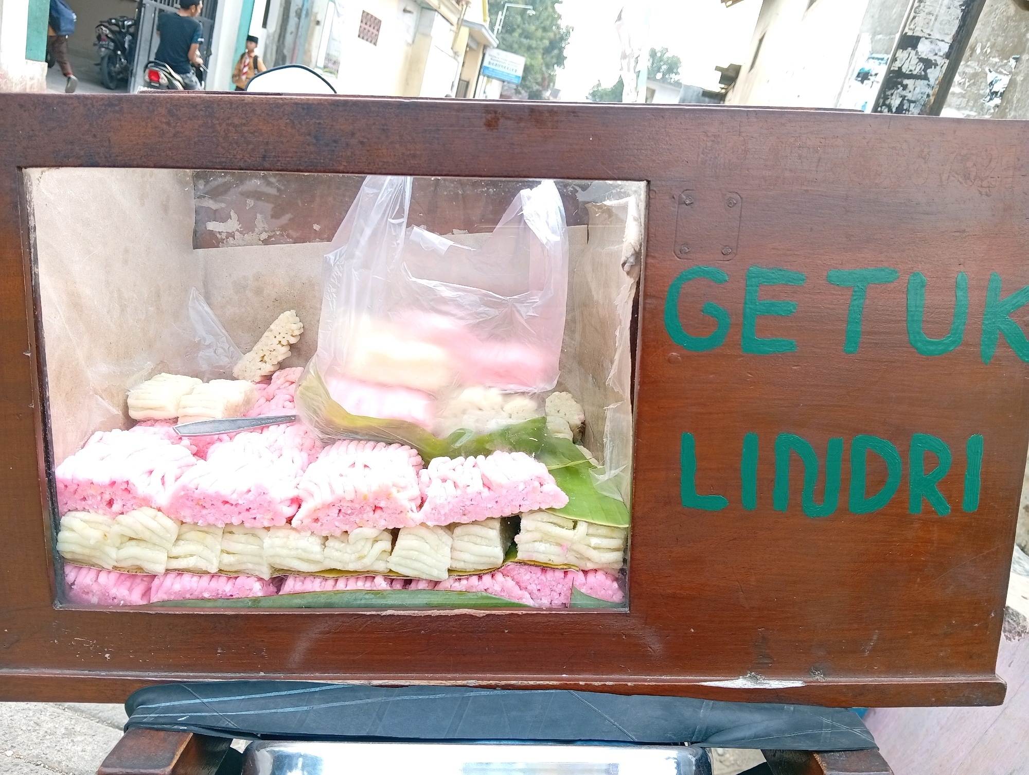 This is a medium shot, eye-level, outdoor photograph taken during the day in Kelutan, Indonesia. The image focuses on a wooden cart displaying food items. The cart has a glass-fronted display case filled with rows of pale pink and white puffed snacks, possibly a type of crackers or candy. Some of the snacks are wrapped in plastic. The exterior of the cart is made of dark brown wood. Green painted text on the right side of the cart reads "GETUK LINDRI". In the background, a person is seen riding a motorcycle and another person stands in front of a building. The environment appears to be a street scene with buildings and a blue sky visible.