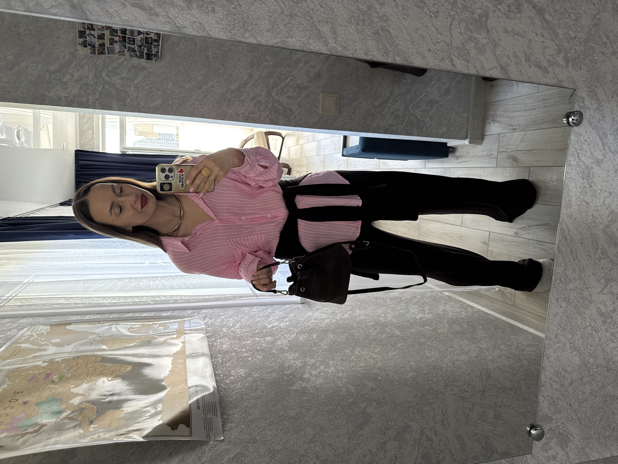 A woman takes a mirror selfie indoors. She has long brown hair, wears a pink and white striped button-down shirt, black pants, and a dark black garment, possibly a sweater, tied around her waist. She accessorizes with a gold-toned choker necklace, a gold ring on her right hand, and carries a small, dark brown shoulder bag. Her lips are covered in dark pink lipstick, and her eyes are closed. She holds a gold/beige-cased smartphone in her right hand to capture the image.
The phone case displays Cyrillic text: "Я выбрала свою любовь" (I chose my love), accompanied by an apple logo.
The room reflected in the mirror features textured grey wallpaper and light wood-patterned flooring. Bright daylight illuminates the scene through large windows fitted with white horizontal blinds. Visible details in the background include a collage of small photographs taped to the wall above the mirror, a white light switch on the wall, and glimpses of furniture such as a dark blue upholstered seat and a wooden chair. A large map, encased in clear plastic and featuring Cyrillic place names like "РОССИЯ" (Russia), is laid out near the window. The mirror is secured with visible silver-toned circular mounts. The setting is a contemporary interior, likely in Sochi, Russia.