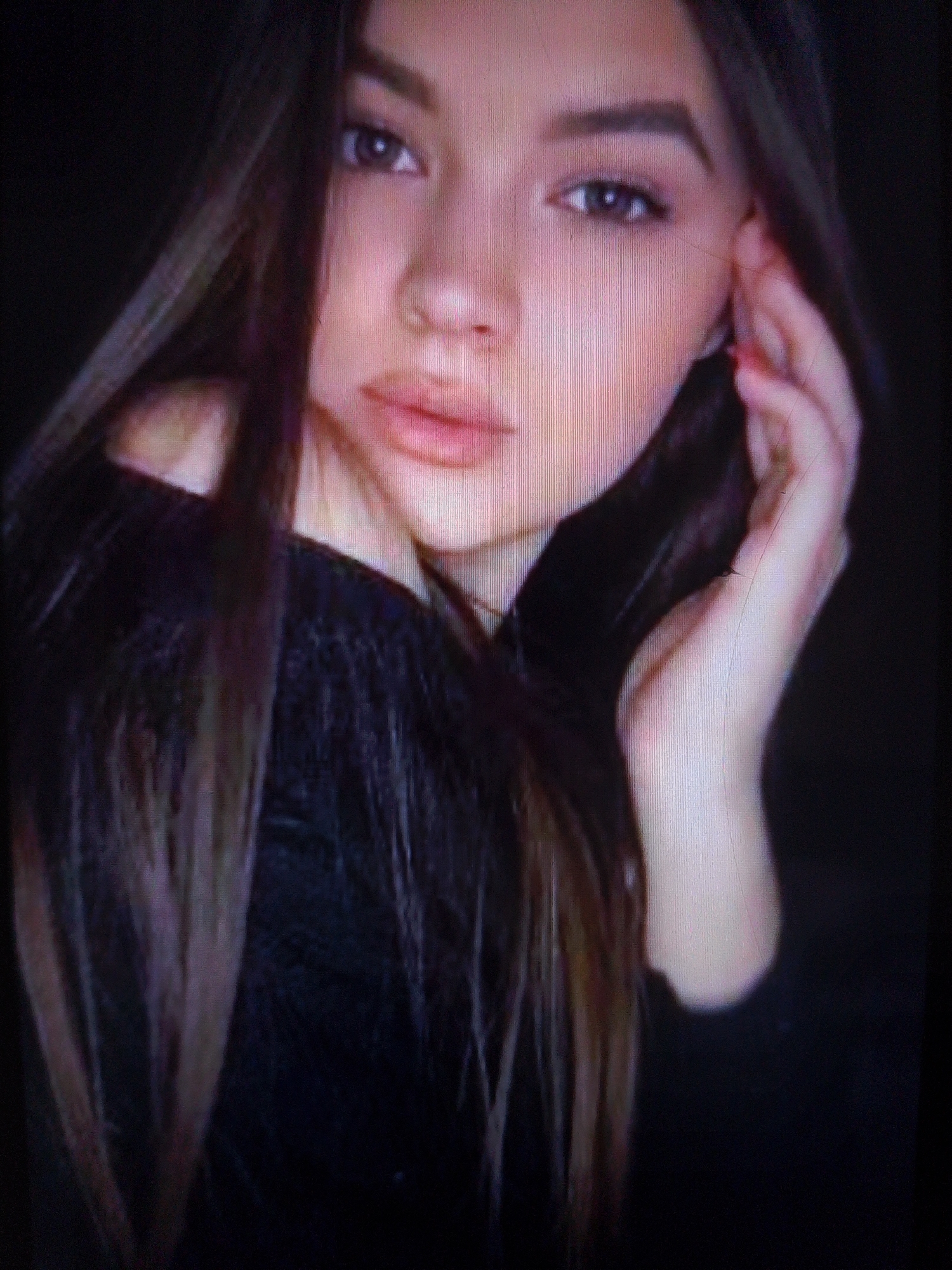 A close-up, low-resolution, indoor portrait shows a young woman with long, dark brown hair and fair skin. She is looking at the camera, and her left hand is raised to her ear, with her fingers touching the side of her head. She is wearing a dark, textured top. The background is dark and out of focus. The image is grainy and appears to be taken with poor lighting. The location in the city of Dobra Krynytsia, Ukraine is not discernible from the provided image.