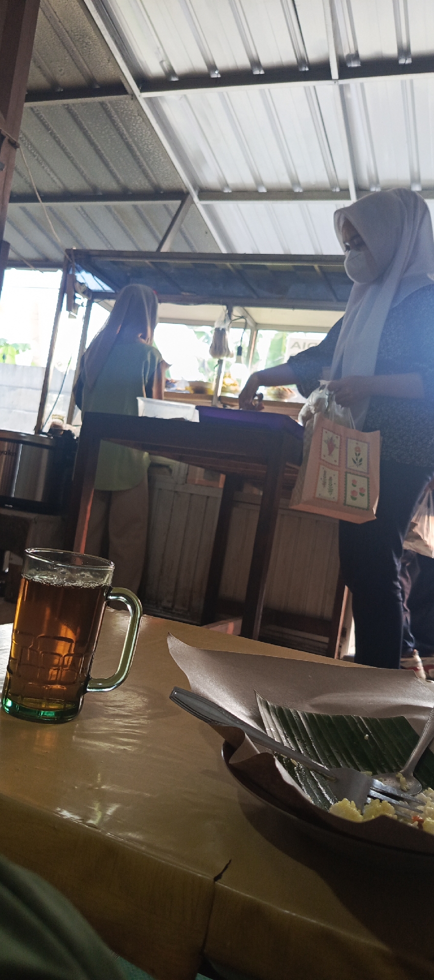 A low-angle shot captures a table in what appears to be an outdoor or semi-open-air food stall or restaurant in Sukaresmi, Indonesia. In the foreground, a glass of iced tea with a green tint and a silver handle sits on a yellowish-brown wooden table. Next to the tea is a plate with food, wrapped in brown paper and a green banana leaf, with a fork resting on top. In the background, two women are visible behind a counter. The woman on the right wears a light blue hijab and a patterned dark blue top, holding a patterned shopping bag. She is wearing a white face mask. The woman on the left wears a light brown hijab and a green shirt, and she appears to be attending to food preparation. The environment suggests a casual dining setting with a corrugated metal roof supported by wooden beams. The lighting is natural, coming from behind the counter.