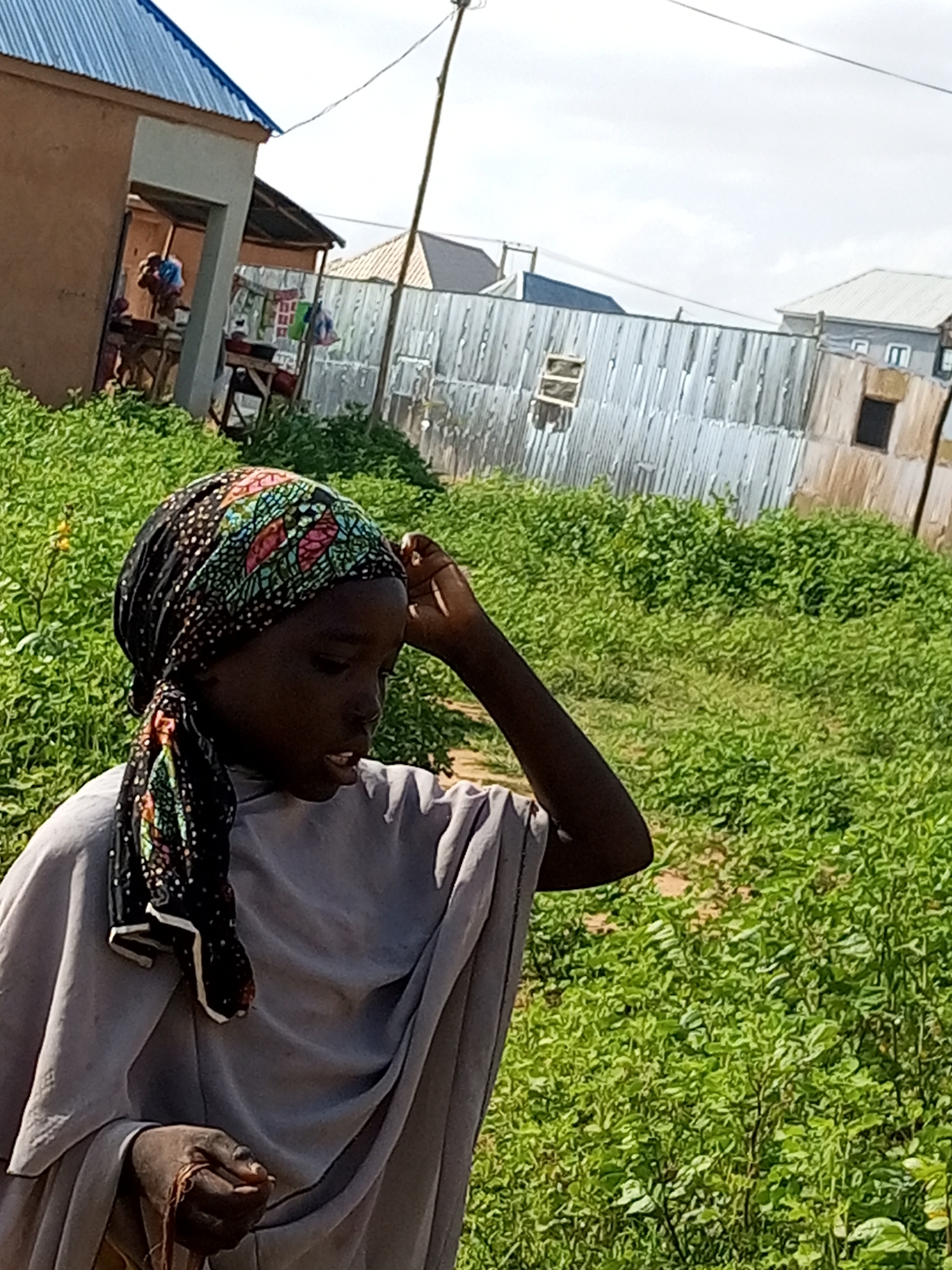 In Tempure, Nigeria, a young girl wearing a headscarf and a gray garment stands outdoors. Her headscarf is black with a colorful floral pattern. She is looking down and to her right, with her right hand touching her head. Her left hand is also visible, holding some thin brown material. The background consists of lush green vegetation, a corrugated metal fence, and a building with a blue metal roof. The sky is partly cloudy.