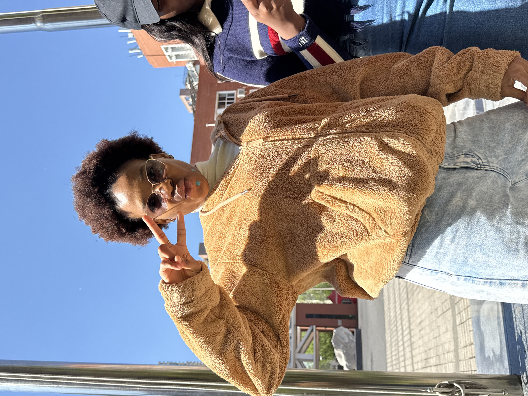 Two individuals are pictured in an outdoor setting, likely in the Daxing District, China based on visual cues. One individual is wearing a brown, fuzzy jacket and light blue jeans, making a peace sign with their hand. They are wearing sunglasses and have short, curly hair. A second individual is partially visible, wearing a dark-colored cap, a blue and white striped sweater with red accents, and dark-colored jeans. The scene is set against a backdrop of a clear, blue sky and a building with brick facade and windows. A silver metal railing is visible in the foreground.