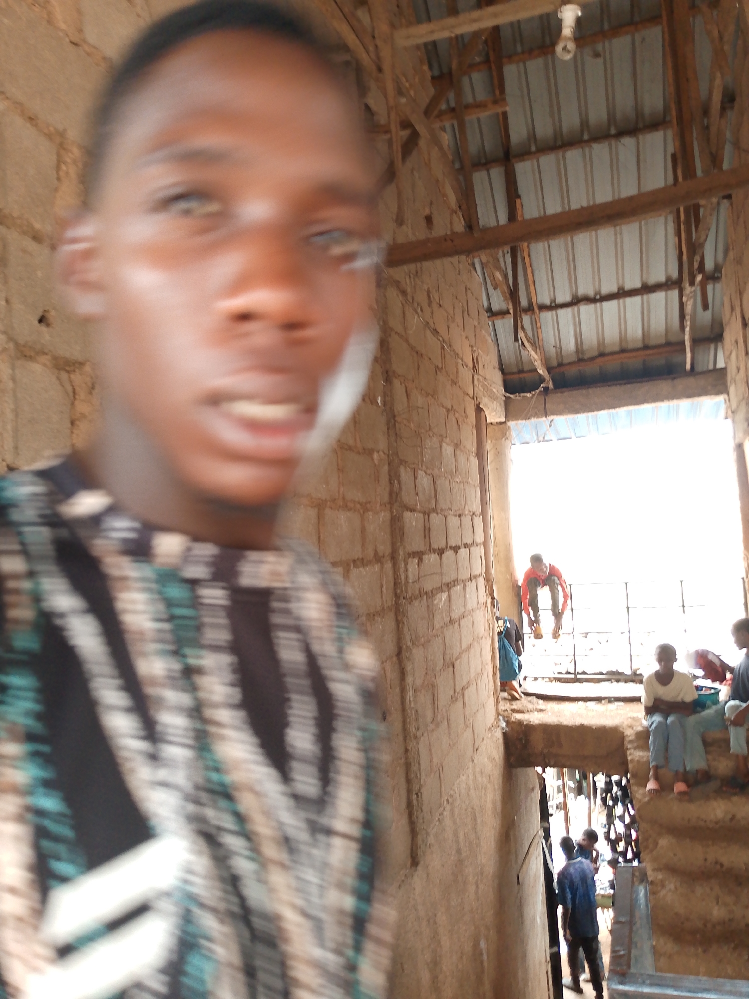 The image presents an out-of-focus individual in the foreground, with a blurred expression and attire featuring a pattern of black, white, and teal. Behind them, a hallway structure with brick walls and a corrugated metal roof is visible. Natural light floods the background, highlighting figures sitting and standing near an opening. A person in an orange shirt is positioned above the others, appearing to interact with a barrier or fence. Below the opening, more people are walking down stairs, suggesting a multi-level structure. The location is identified as Unknown, Unknown.