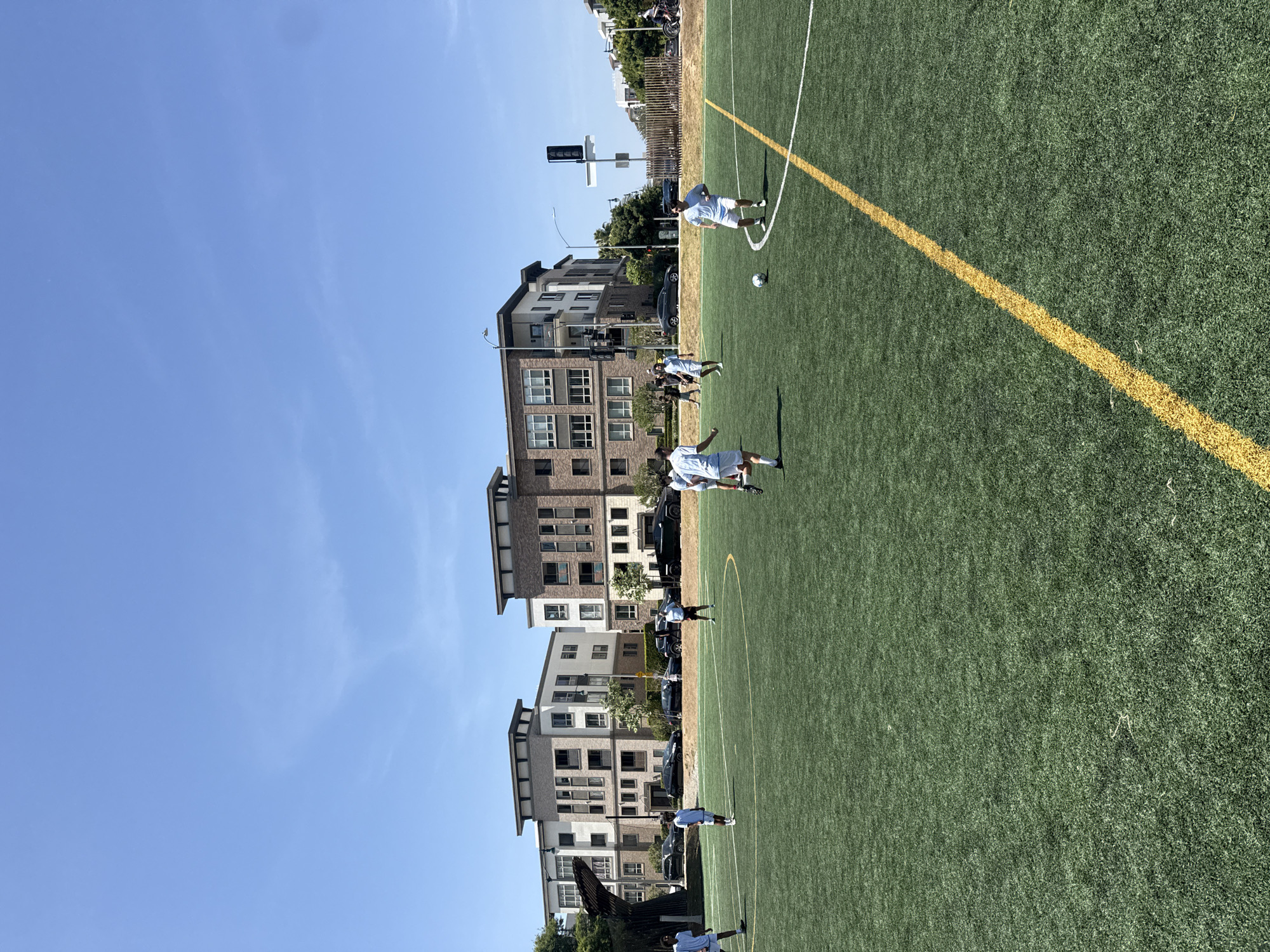 A soccer game is underway on an artificial turf field adjacent to a row of multi-story residential buildings under a clear blue sky. The yellow lines mark the boundaries of the field. Several players, dressed in white jerseys and shorts, are scattered across the field. The buildings are brown and white, with a mix of brick and siding exteriors. Cars are parked along the street in front of the buildings. The location is consistent with an urban or suburban area. The city of Los Angeles cannot be positively determined.