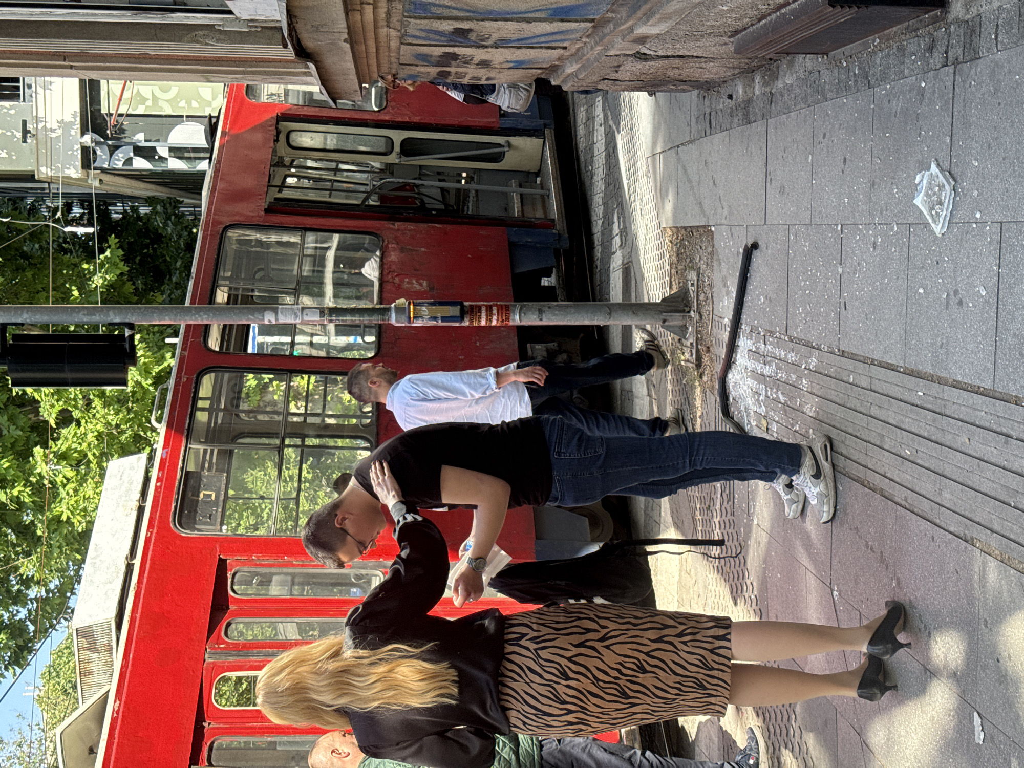 On a sunny street in Belgrade, Serbia, a group of people are standing next to a red tram. A woman with long blonde hair, wearing a black jacket and zebra print skirt, is standing with her back to the camera. Next to her is a man in a black t-shirt, blue jeans, and white sneakers, appearing to adjust his glasses. Another man in a white shirt and dark pants stands behind him. The tram, with its faded red paint and multiple windows, is positioned next to the sidewalk. A traffic light pole is present on the left side of the frame, obscured by the foliage. On the sidewalk, broken glass pieces are scattered, and a black cane lies on the ground. The background includes buildings and trees, contributing to an urban landscape.
