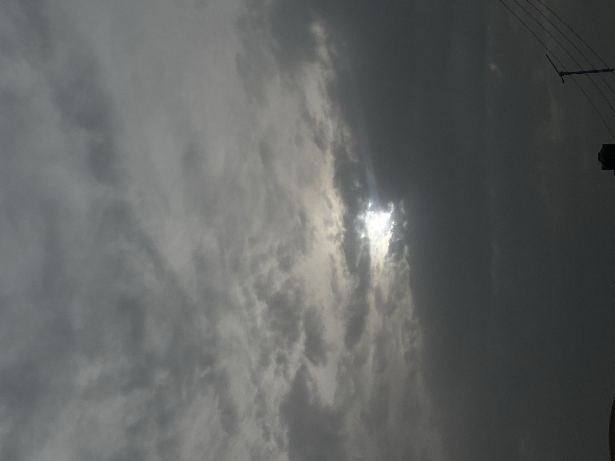 The image shows a cloudy sky in Barden Yana, Nigeria. The sun is partially obscured by thick, gray clouds, creating a diffused light. In the upper right corner, there is a utility pole with wires extending from it. The environment suggests overcast conditions. There are no people, subjects, objects, actions, or interactions visible beyond the described elements.