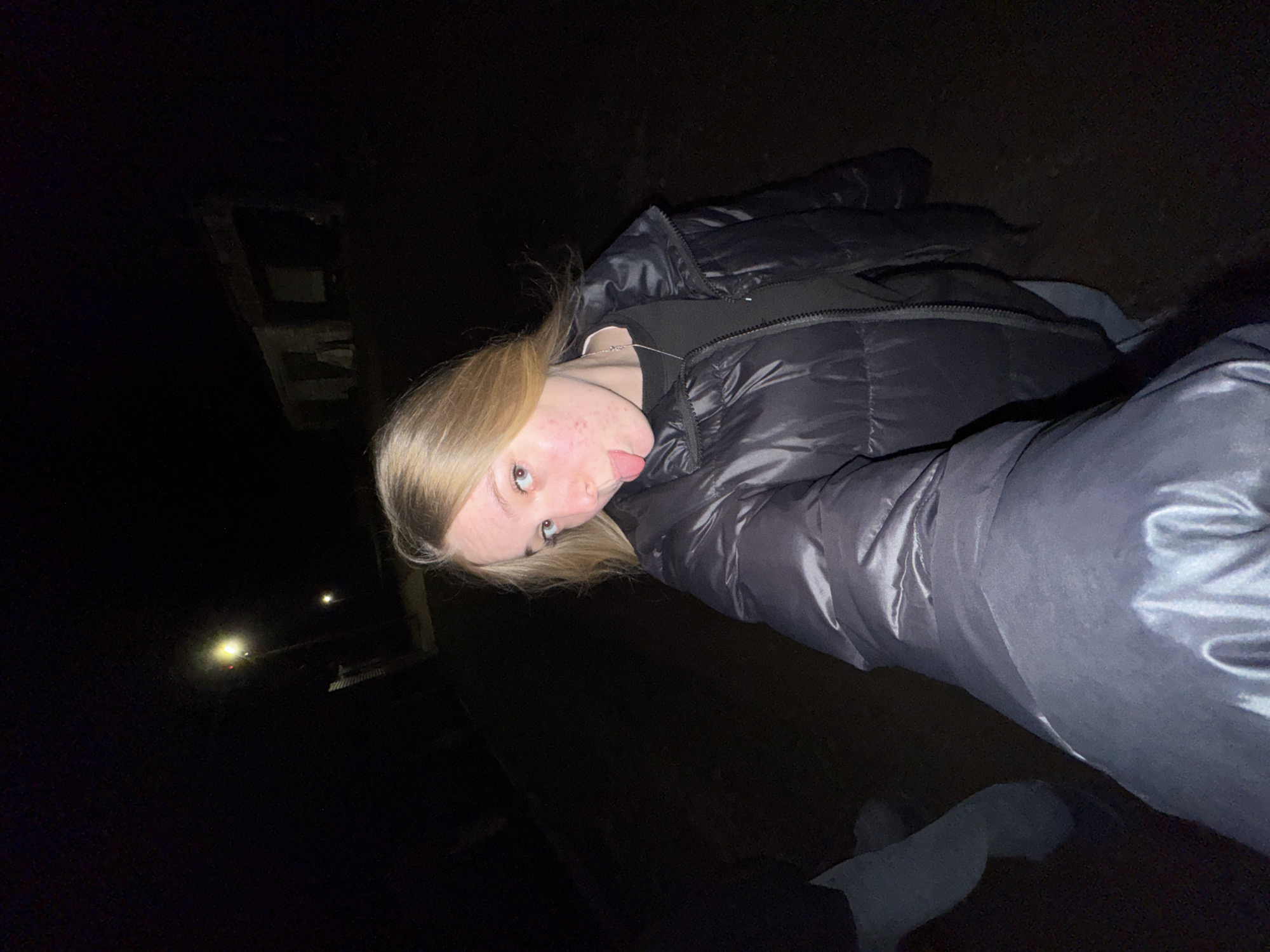 A person with blonde hair is lying on the ground outside at night. The person is wearing a black puffy jacket. Their tongue is sticking out. The location may be near a building with a lit window. Zhigalki, Russia, cannot be confirmed without additional information.