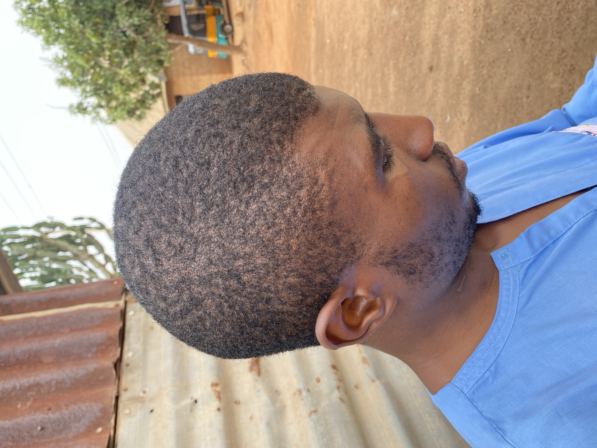 A close-up, profile view photograph depicts a dark-skinned man with a closely-cropped, dark, textured hairstyle, a trimmed beard, and wearing a light blue shirt. He is positioned outdoors. In the background, a tree with green foliage and a partially visible structure are seen. To the left, a corrugated metal sheet is visible. The location is Kuru Karama, Nigeria.