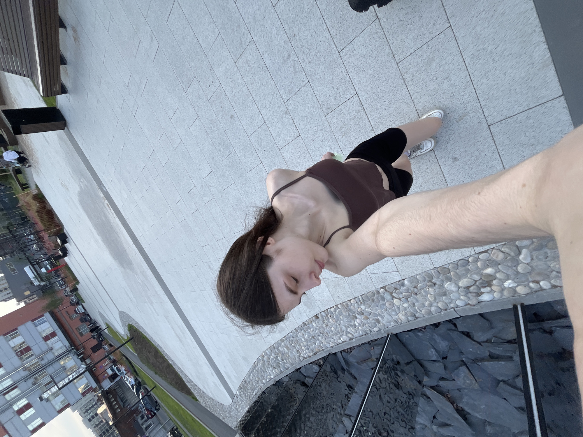 A young woman with long brown hair, wearing a brown spaghetti-strap tank top, black shorts, and white sneakers, is captured in a high-angle view, holding the camera with her left arm partially visible in the foreground. She is looking downwards and to her left. To her immediate left is a curved decorative water feature with an edge composed of numerous small, light-colored rounded pebbles. Below this, dark, irregularly shaped stones line the bottom of a shallow basin, with the water surface reflecting the environment. The ground she stands on is paved with light grey rectangular slabs. In the mid-ground to the right, a person in a white top pushes a light-colored stroller or cart near a wooden slatted bench and a dark trash can. Further in the background, an urban street with parked cars runs parallel to a grassy strip. Beyond the street, multi-story buildings are visible, including one with a grey facade and red window accents, and a reddish-brown structure. The scene depicts a modern urban public space, specifically the revitalized **Karpovka Embankment** in Saint Petersburg, Russia. The lighting is natural daylight, suggesting an overcast sky or late afternoon.