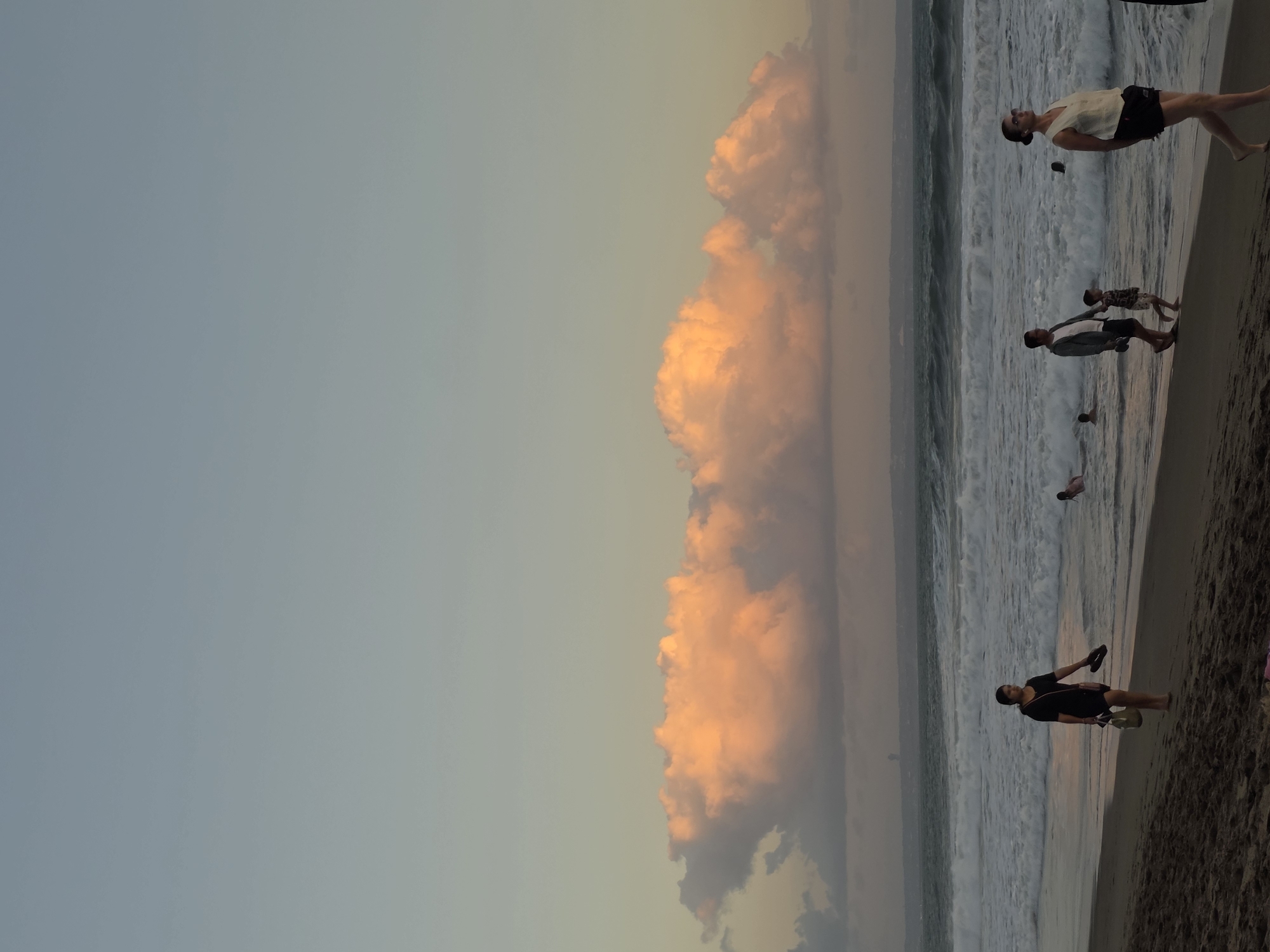 The image captures a serene sunset scene on a beach in North Kuta, Indonesia. The sky, transitioning from a pale blue at the top to a soft orange and pink hue near the horizon, is adorned with large, fluffy clouds illuminated by the setting sun. The ocean waves are active, creating a white foamy texture as they meet the sandy shore.
Several individuals are present on the beach. In the foreground, a woman in a black dress walks along the waterline, carrying a bag and flip-flops. Further down the beach, a couple walks hand-in-hand with a young child. To the right, a woman in a white top and black shorts walks towards the viewer. Other people are visible in the shallow water, some swimming and others wading. In the distance, a faint silhouette of a city skyline or industrial structure is visible across the water. The sand is dark and appears wet near the water's edge.
