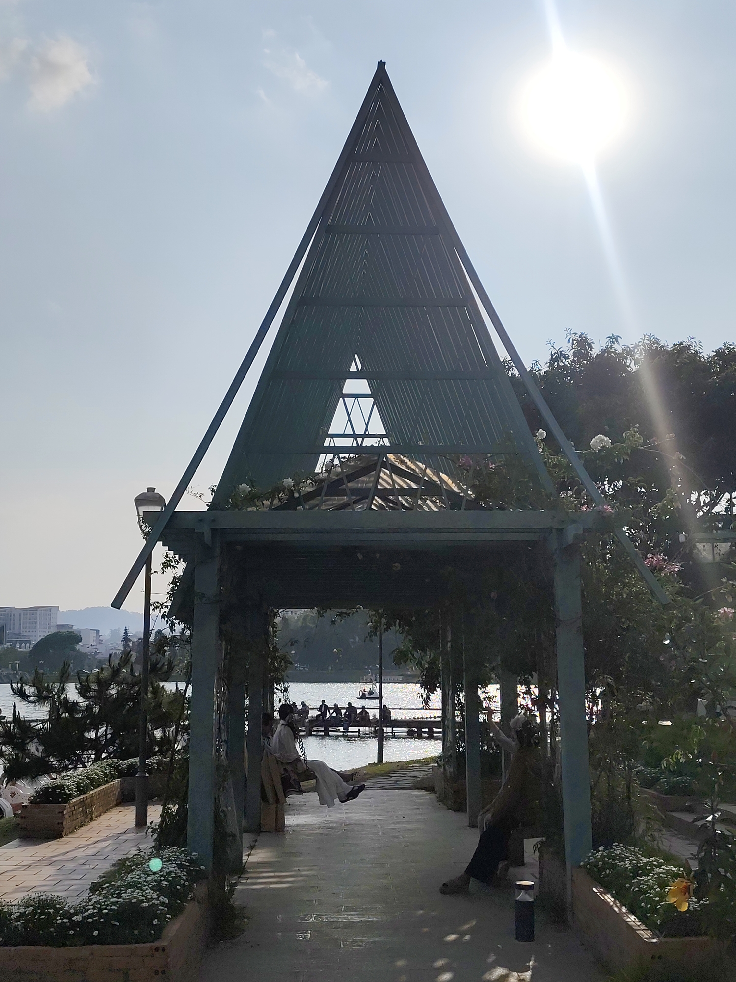 A daytime shot shows an open-air structure in Da Lat, Vietnam, near a lake. The structure is a tall, pyramid-like pergola with a metal frame and a triangular opening at the top. Vines and flowers are growing on the pergola. Inside the pergola, people are sitting on benches. Outside the structure, planters of flowers and shrubs line the pathway. A lamppost stands near the pergola. In the background is a lake, with people in boats and a distant city skyline. The sun is visible, causing a bright flare in the upper right corner.