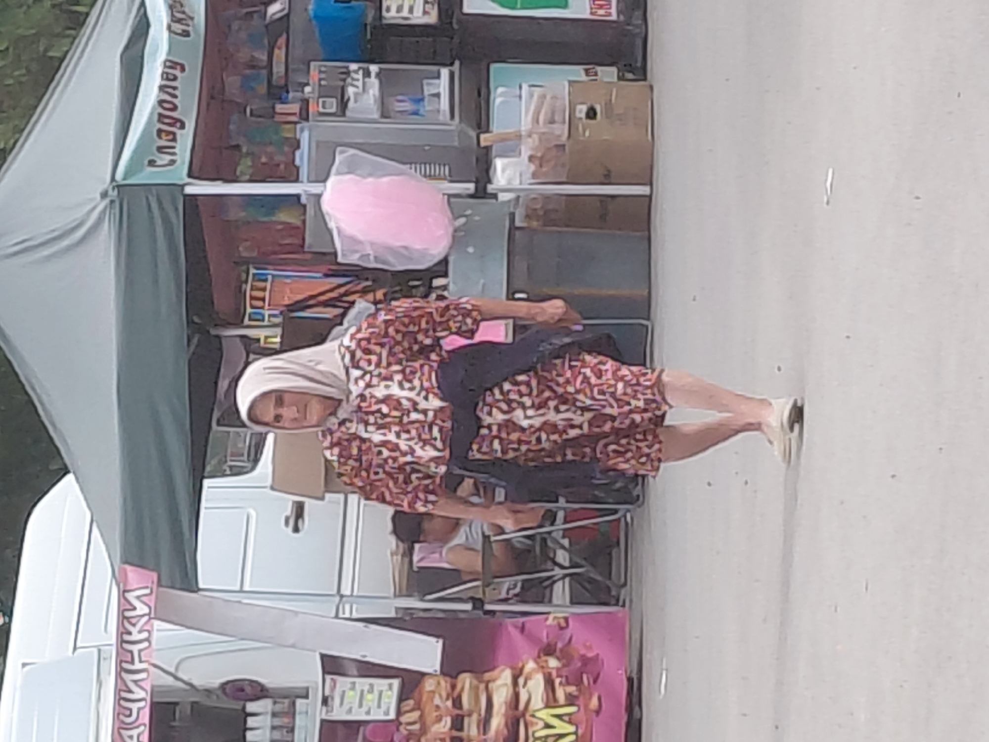 The image depicts an outdoor market scene in Kostievo, Bulgaria, featuring two vendor tents and two individuals. In the foreground, an older woman walks from right to left across a paved surface. She wears a light-colored headscarf, a short-sleeved dress with a multi-colored abstract pattern (red, brown, white), and light-colored flat shoes. A dark blue or black garment is tied around her waist. Her expression is neutral.
Behind her, to the left, is a green canopy tent with a pink banner partially displaying the Cyrillic text "ЧИНКИ" (likely part of a food item name like pancakes). A large pink sign with an image of stacked pancakes is visible next to this tent. A white van or food truck is parked behind the tent. Underneath this tent, a person with short hair, wearing a light-colored sleeveless top, sits in a metal folding chair, looking down at something in their lap.
To the right, another green canopy tent has a light blue banner displaying "Сладолед" (Ice Cream) in Cyrillic. This tent features a large, transparent bag of pink cotton candy hanging prominently. Various market goods, including colorful packaged items and what appears to be a refrigeration unit, are visible inside the stall. The ground is a light-colored, possibly concrete or asphalt, surface with minor debris. Green foliage is discernible in the distant background above the tents.