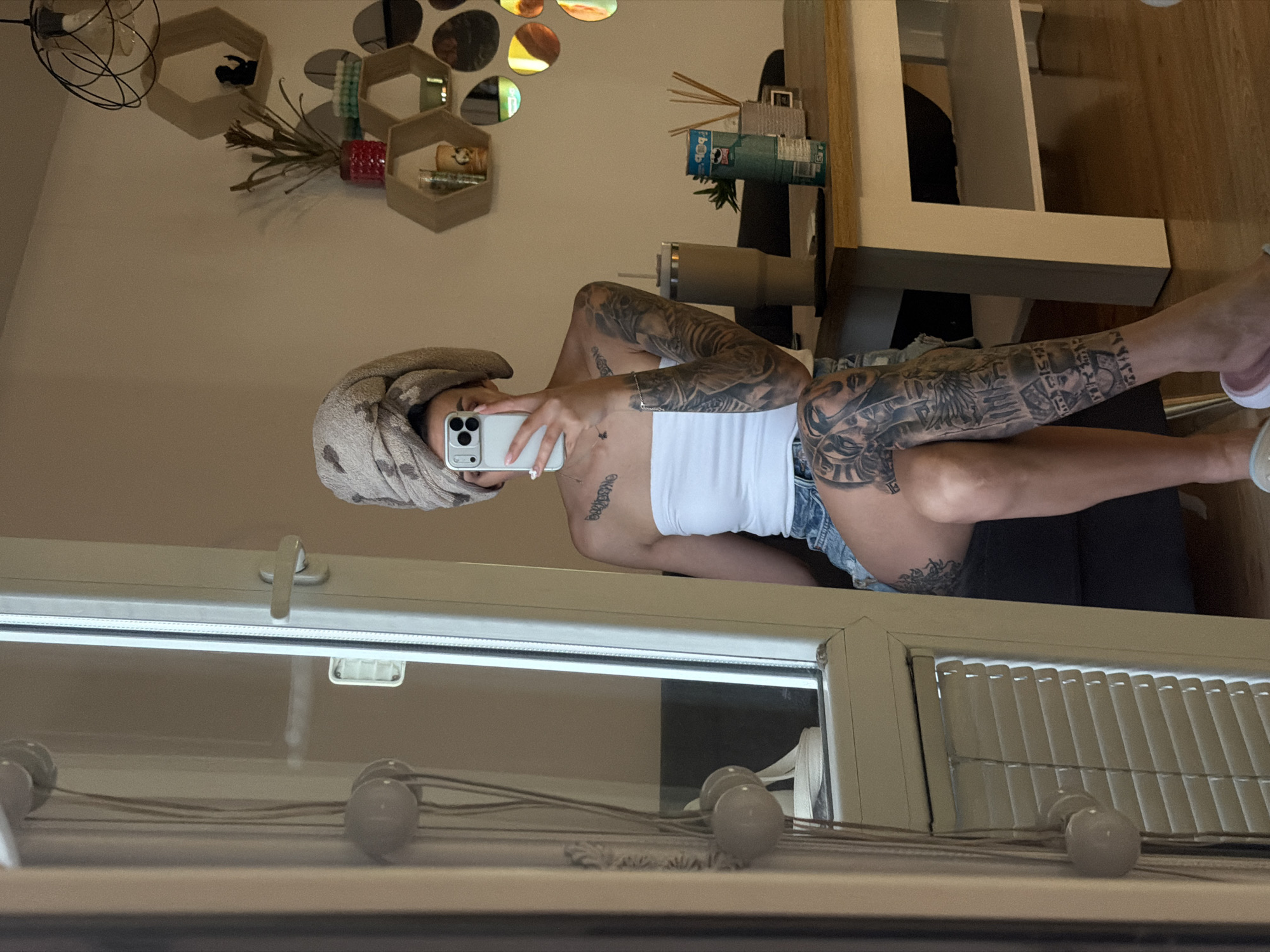 The depicted scene is an indoor setting within a residence, identified as being in Plovdiv, Bulgaria. An adult individual is captured in a reflection, likely a large window or mirror, taking a selfie. The person's head is wrapped in a light-colored, patterned towel. A light-colored smartphone, held in the right hand, obscures the majority of the face. The individual wears a white strapless crop top and denim shorts. Extensive dark-toned tattoos are visible on both arms, forming full sleeves on the right arm and partially on the left shoulder and chest, and also cover both legs, with a full sleeve on the right leg and a partial design on the left thigh. The tattoos appear to depict intricate patterns and figural elements. A delicate necklace adorns the neck. Light-colored slides or slippers are partially visible on the feet.
The background shows a light-colored wall adorned with three hexagonal wooden shelves, vertically stacked on the right. A small green plant in a red pot and a dark figurine (possibly a monkey) are placed on the shelves. Above these, a black wire-frame pendant light fixture hangs from the ceiling. Circular mirror decals are arranged decoratively on the wall to the right of the shelves. A light-toned wooden table with a white base occupies the lower right portion of the image. On the table, a large beige insulated tumbler, a green Pringles can, a small plant, and a reed diffuser are visible. A dark-colored chair or sofa is partially visible behind the individual and to the left of the table. The floor is covered with light-toned wooden flooring.
The left side of the reflection prominently displays a white window frame with a handle. Through the lower portion of the window, white horizontal blinds are visible. To the far left, a decorative object featuring multiple light-colored spherical elements connected by cords is partially reflected.