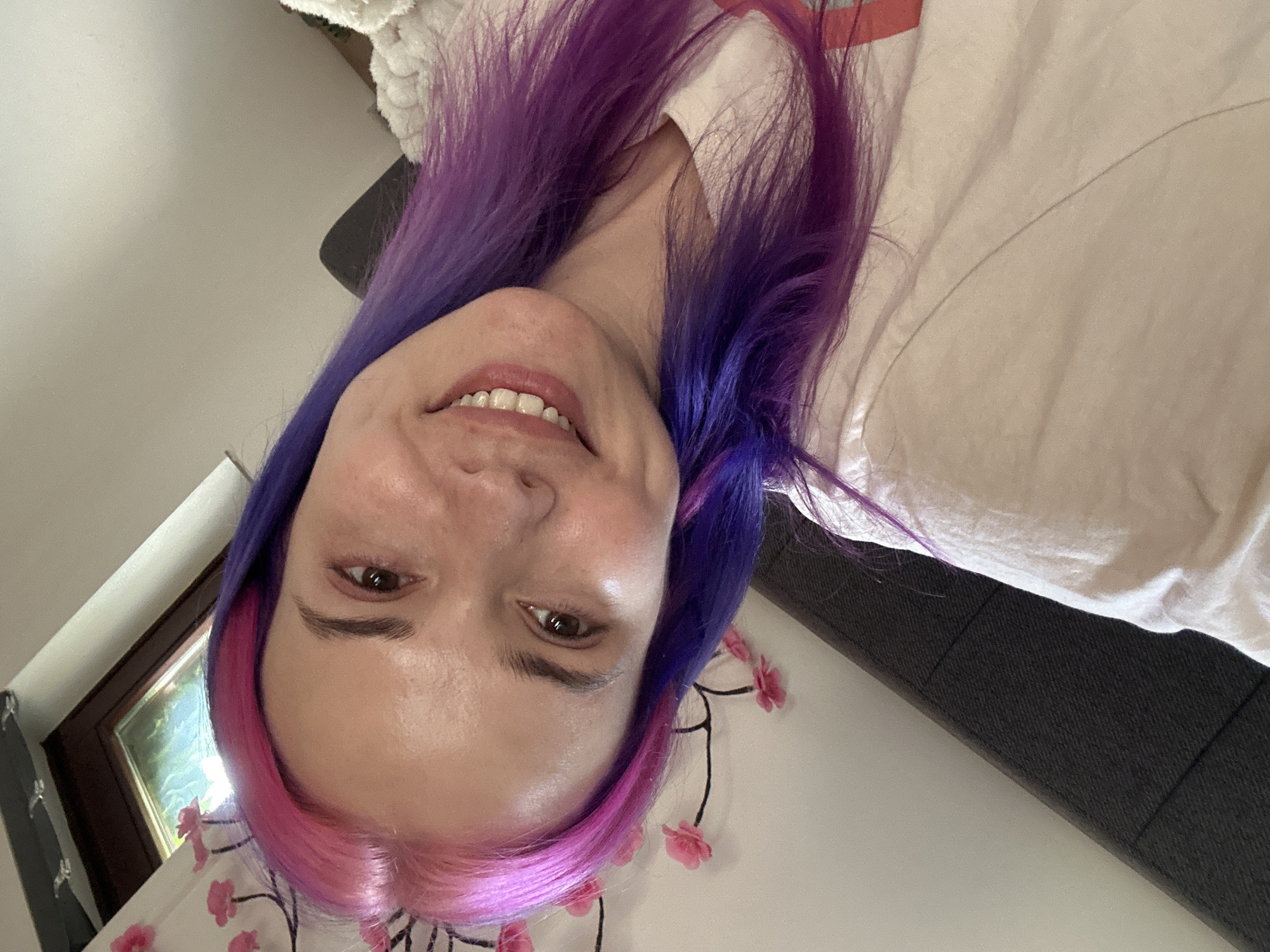 The image shows a woman with dyed hair, the top half pink and the bottom half purple, smiling at the camera. She is wearing a white shirt with a red stripe. A window is visible in the background. A gray couch and a blanket with pink flowers are also visible. The location cannot be determined from the image.